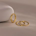 Three thin gold bands from the Simple Ring Set rest on a smooth beige surface, with one stacking ring propped against a curved white object.