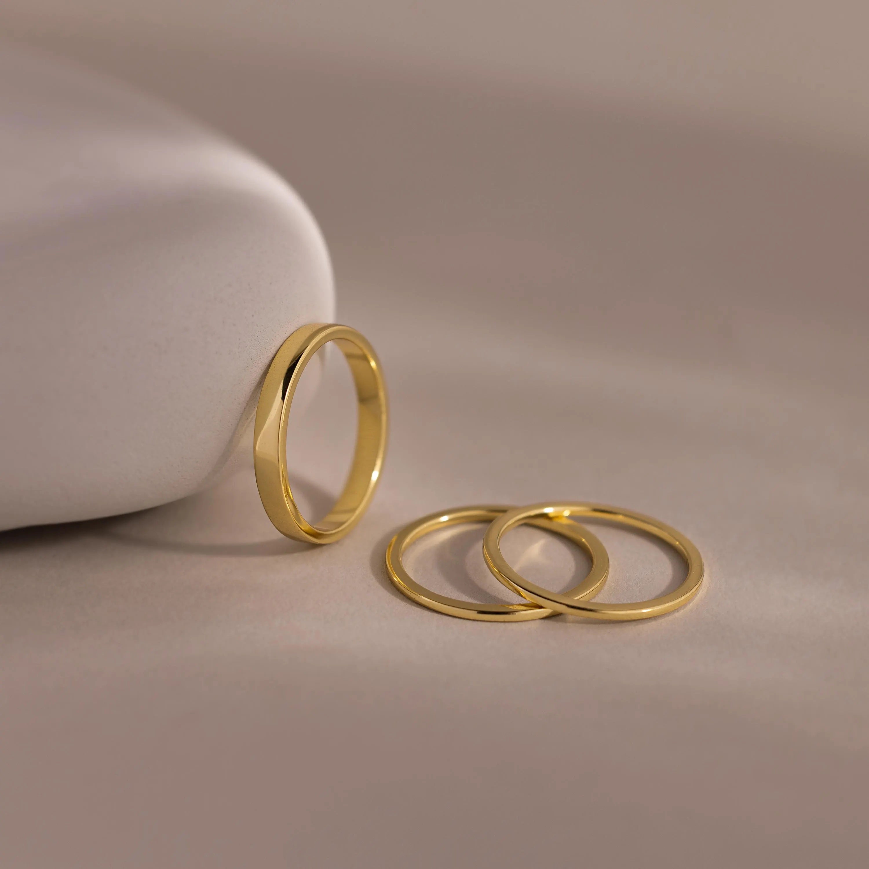 Three thin gold bands from the Simple Ring Set rest on a smooth beige surface, with one stacking ring propped against a curved white object.