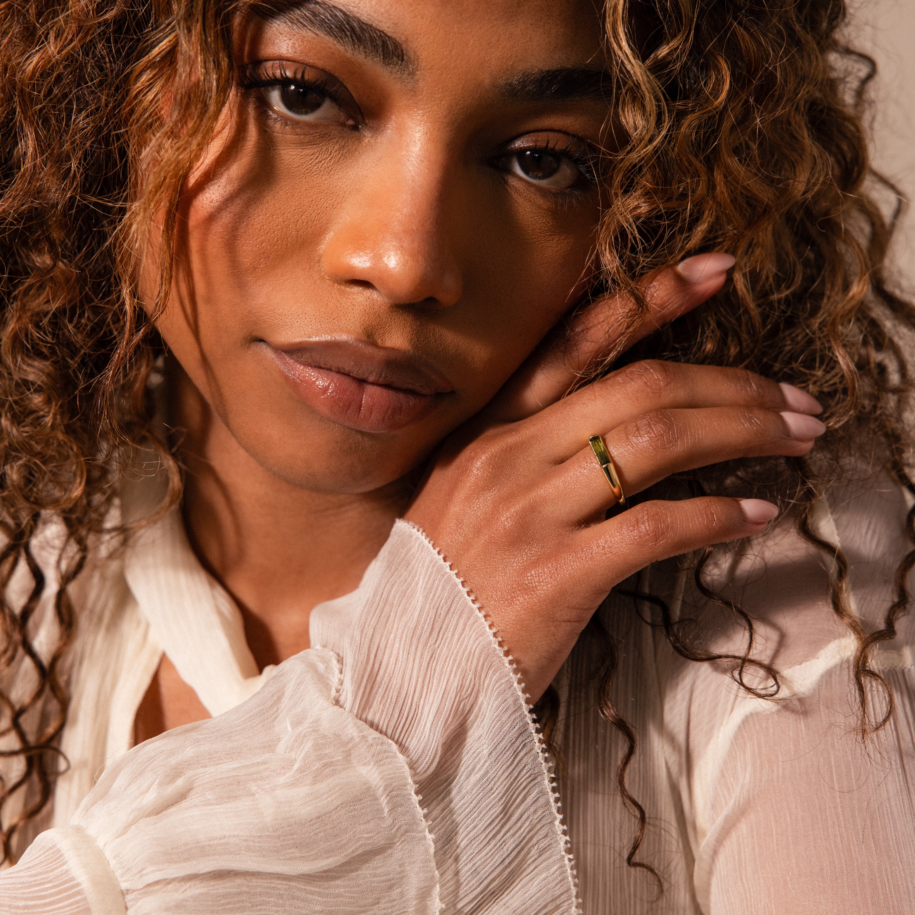 A woman with curly hair in a white blouse touches her face, wearing the Baguette Signet Ring as a modern statement piece while looking at the camera.