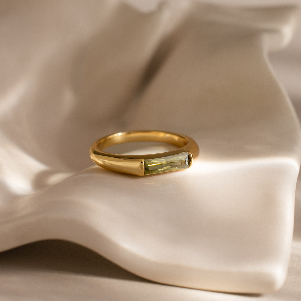 The Baguette Signet Ring in gold, set with a rectangular green gemstone, is showcased on an ivory-colored fabric background.