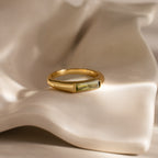 The Baguette Signet Ring in gold, set with a rectangular green gemstone, is showcased on an ivory-colored fabric background.