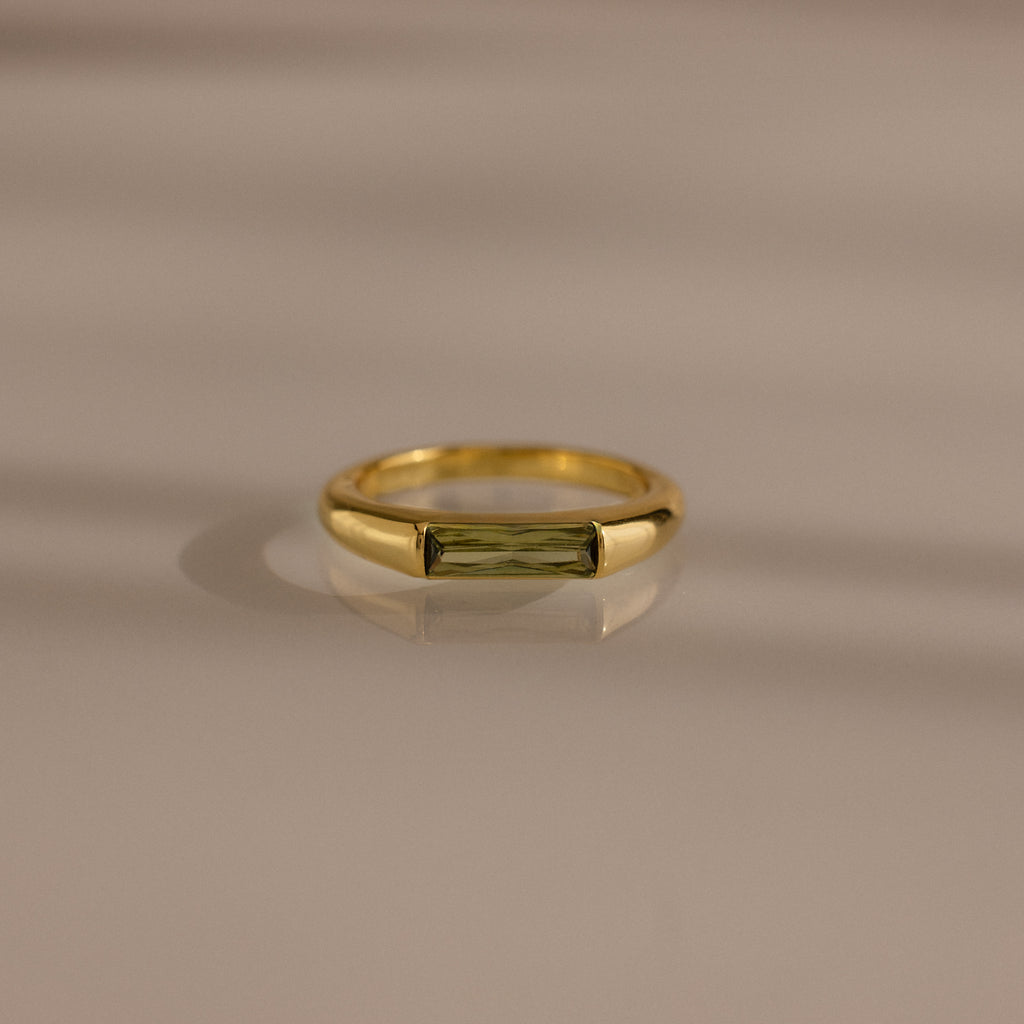 The Peridot Baguette Signet Ring is a modern gold statement ring featuring a rectangular green peridot gemstone in a baguette cut, showcased on a smooth beige surface with soft shadows.