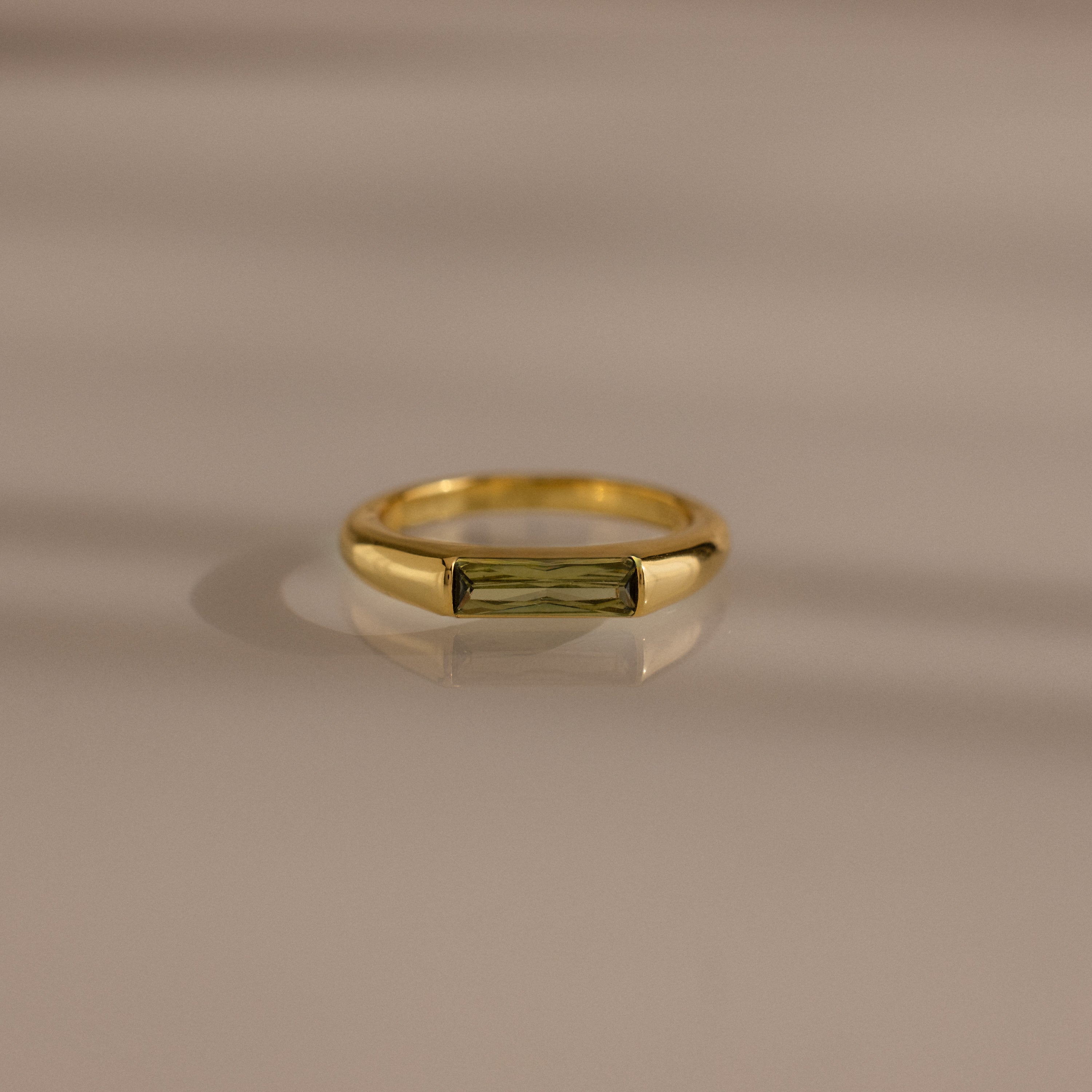 The Peridot Baguette Signet Ring is a modern gold statement ring featuring a rectangular green peridot gemstone in a baguette cut, showcased on a smooth beige surface with soft shadows.