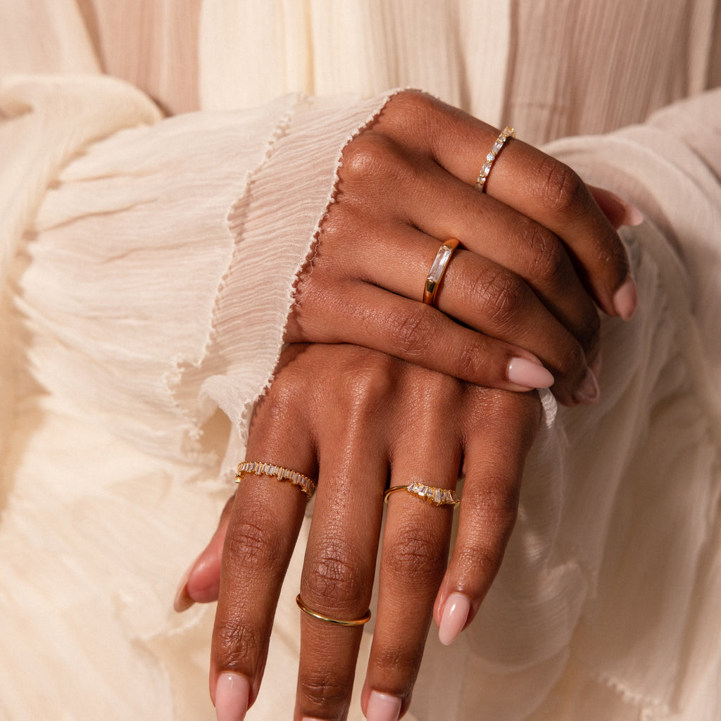 A close-up of manicured hands wearing several delicate gold and diamond rings, including the stackable Art Deco Rhythm Ring set with sparkling baguette diamonds.