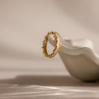 The Art Deco Rhythm Ring, featuring a textured gold design, rests on a smooth white sculptural surface under soft lighting.