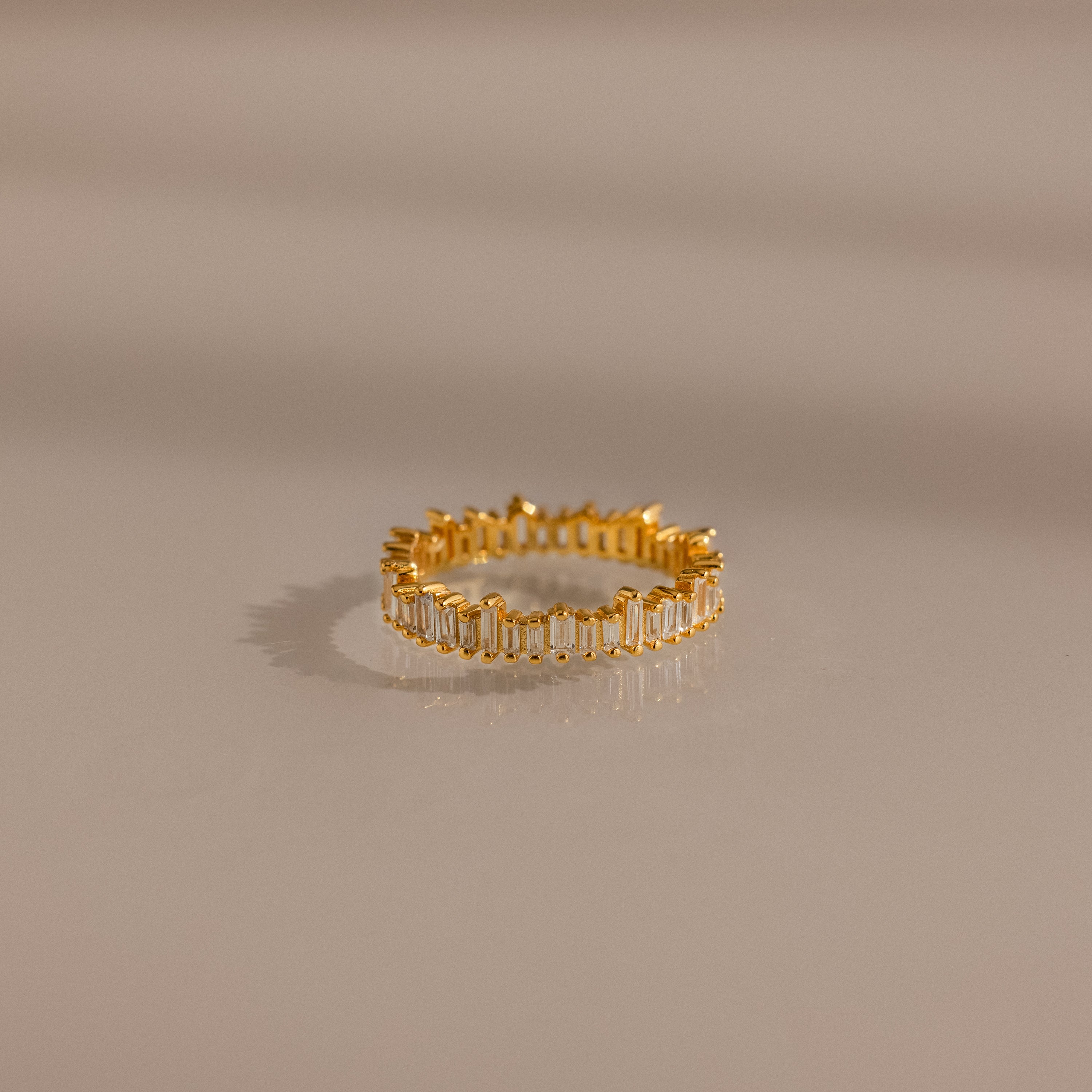 The Art Deco Rhythm Ring features rectangular baguette diamonds set in a continuous gold band, showcased on a smooth beige surface.