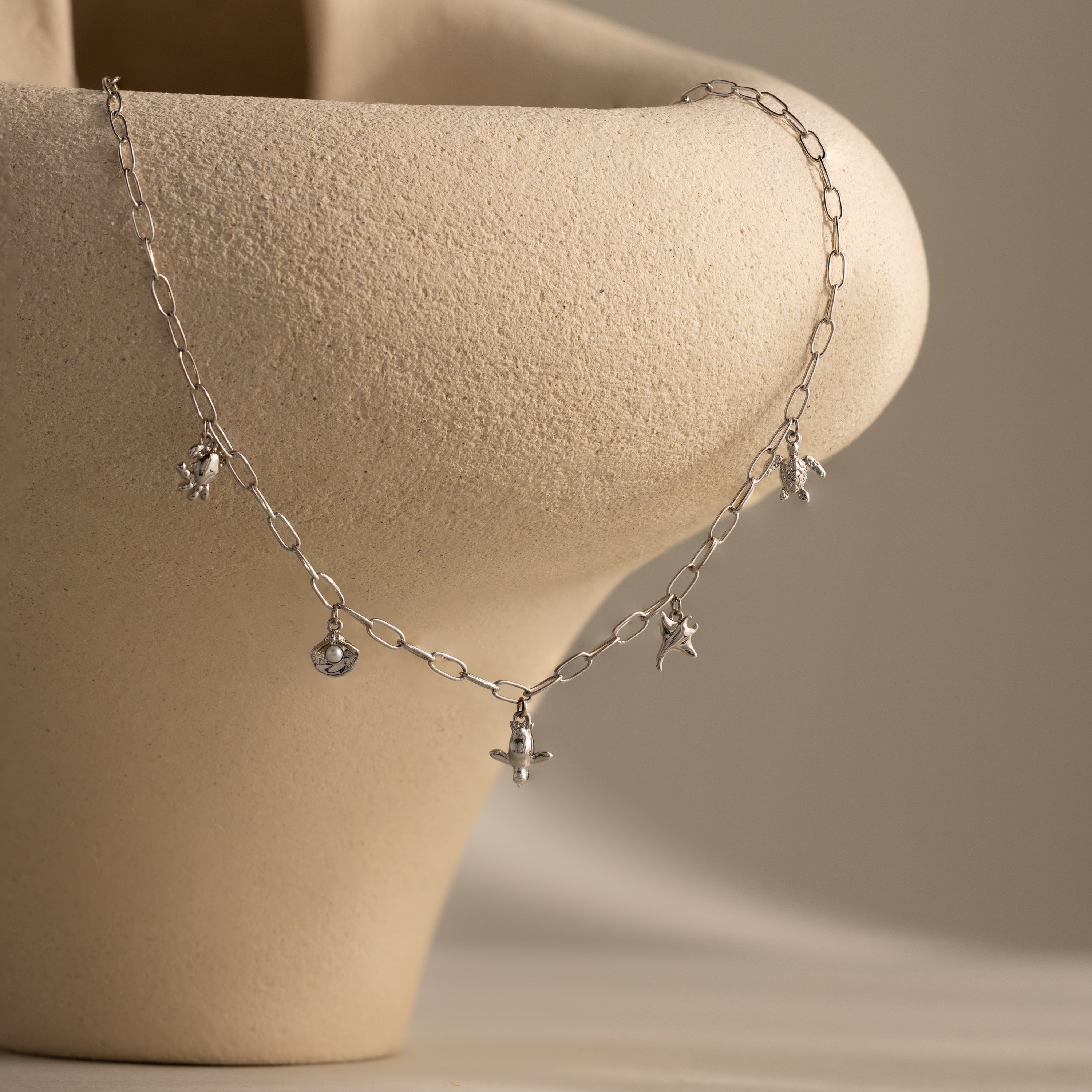 The Seaside Charm Necklace in Silver features star and bead charms, draped over a textured beige ceramic vase—perfect as summer or beach jewelry.
