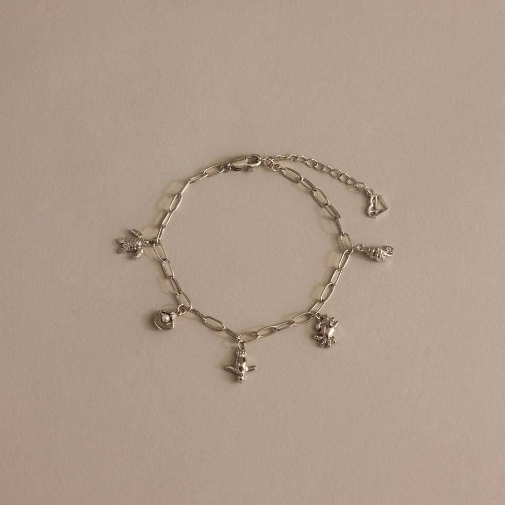 Seaside Charm Bracelet in Silver: Features five ocean-inspired charms—heart, angel, and animal—on a silver chain, displayed against a neutral beige background.