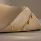 The Seaside Charm Bracelet in 18K Gold features five unique ocean-inspired charms displayed on a curved, textured beige surface.