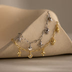 Two Seaside Charm Bracelets, one in silver and one in gold, with ocean-inspired charms, draped over a beige geometric surface with sculpted shapes.