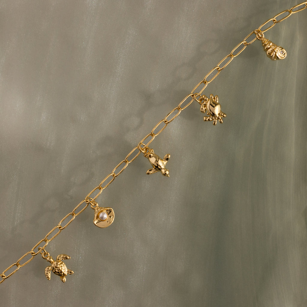 Seaside Charm Bracelet featuring gold chain and ocean-inspired charms—turtle, shell, fish, and seashell—displayed on a gray surface.