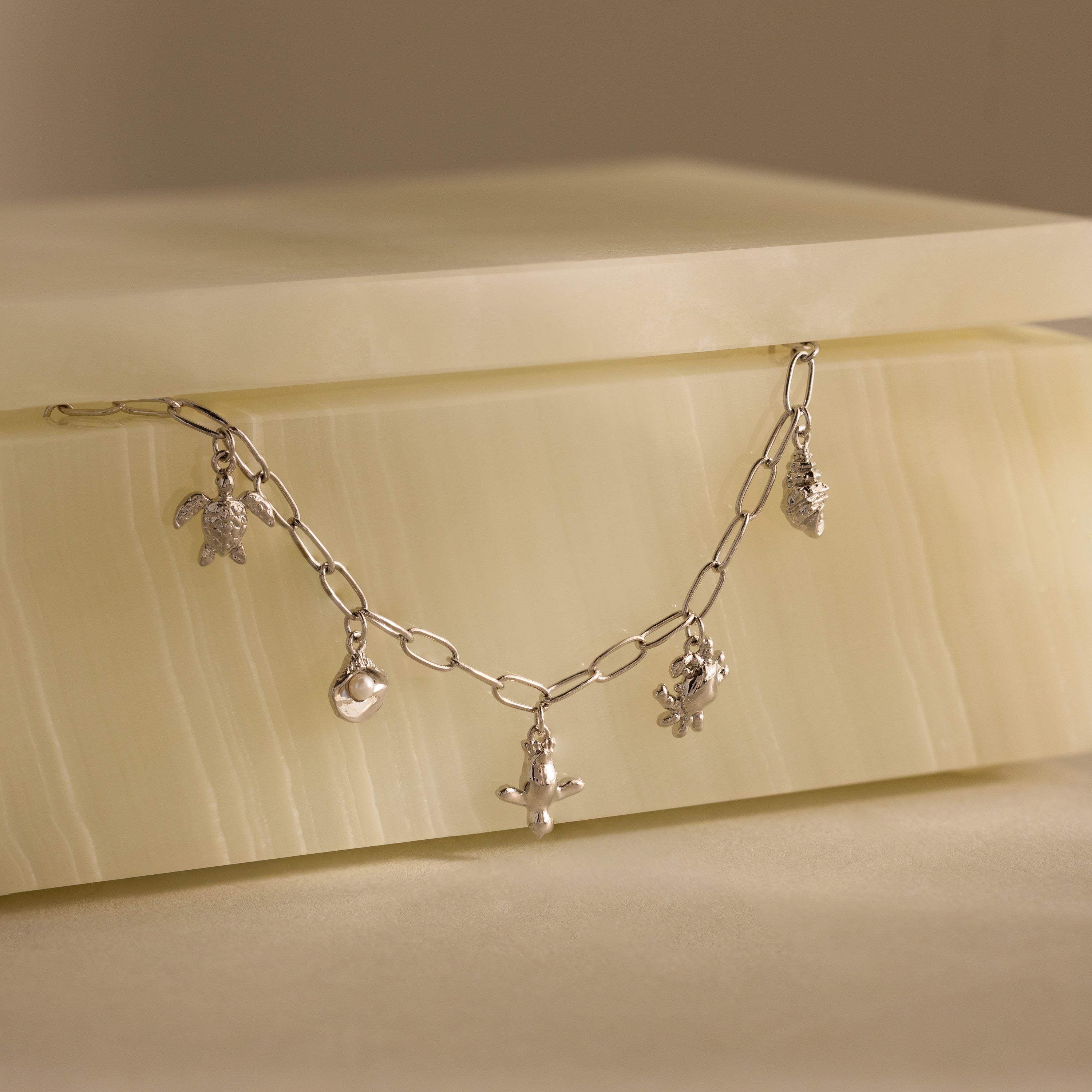 The Seaside Charm Bracelet features turtle, moon, cross, and pineapple ocean-inspired charms displayed on a cream-colored stone box.