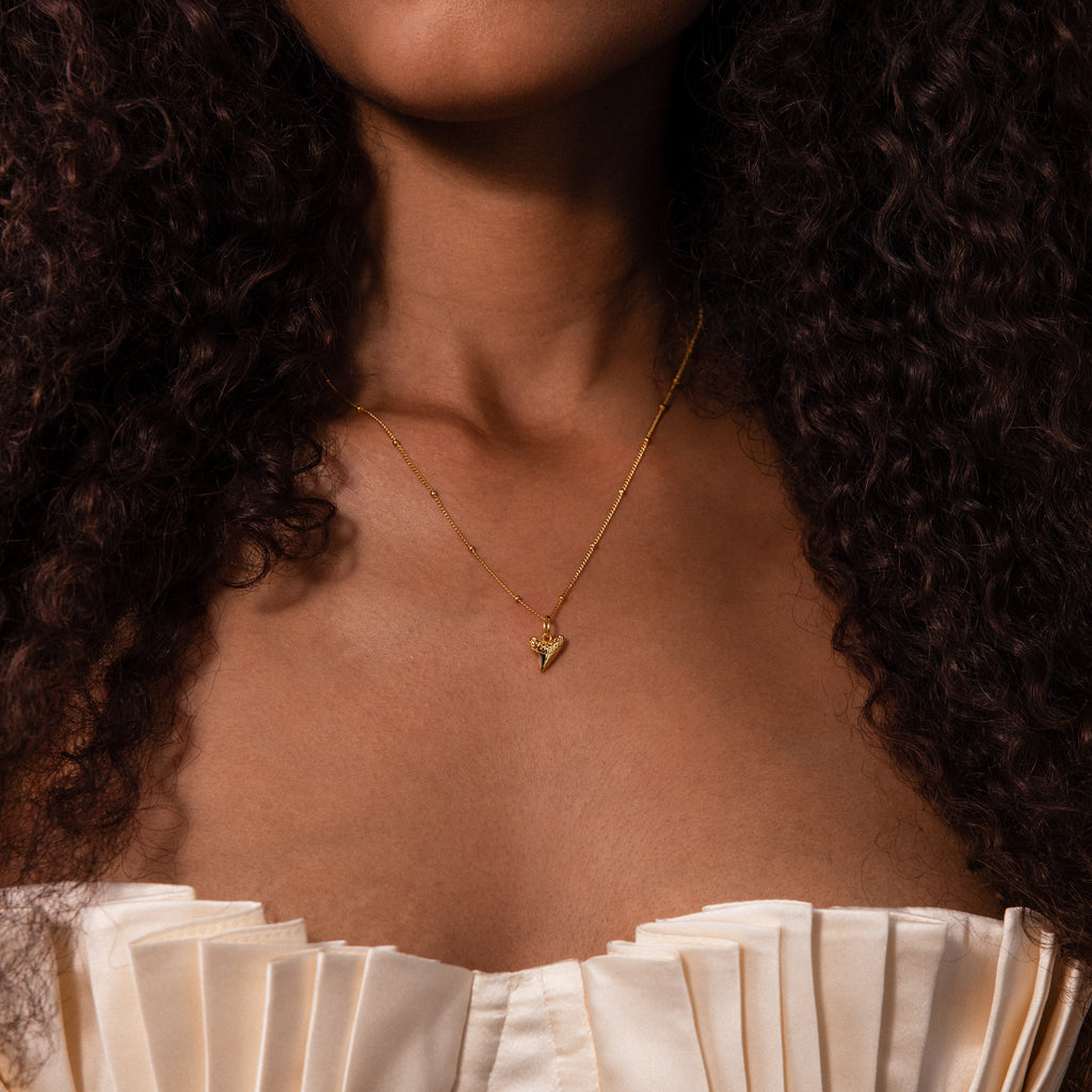 Woman wearing the Shark Tooth Necklace in 18K Gold, featuring an Africa-shaped pendant, paired with a cream pleated off-shoulder top.