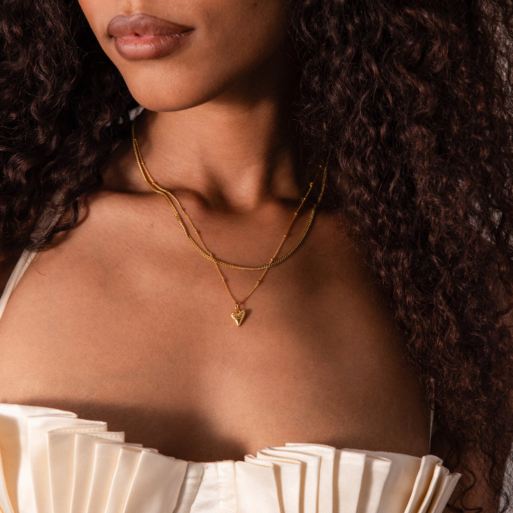 A woman shown from shoulders to lips wears a cream pleated strapless top and the Shark Tooth Necklace in 18K Gold, layered with other gold necklaces.