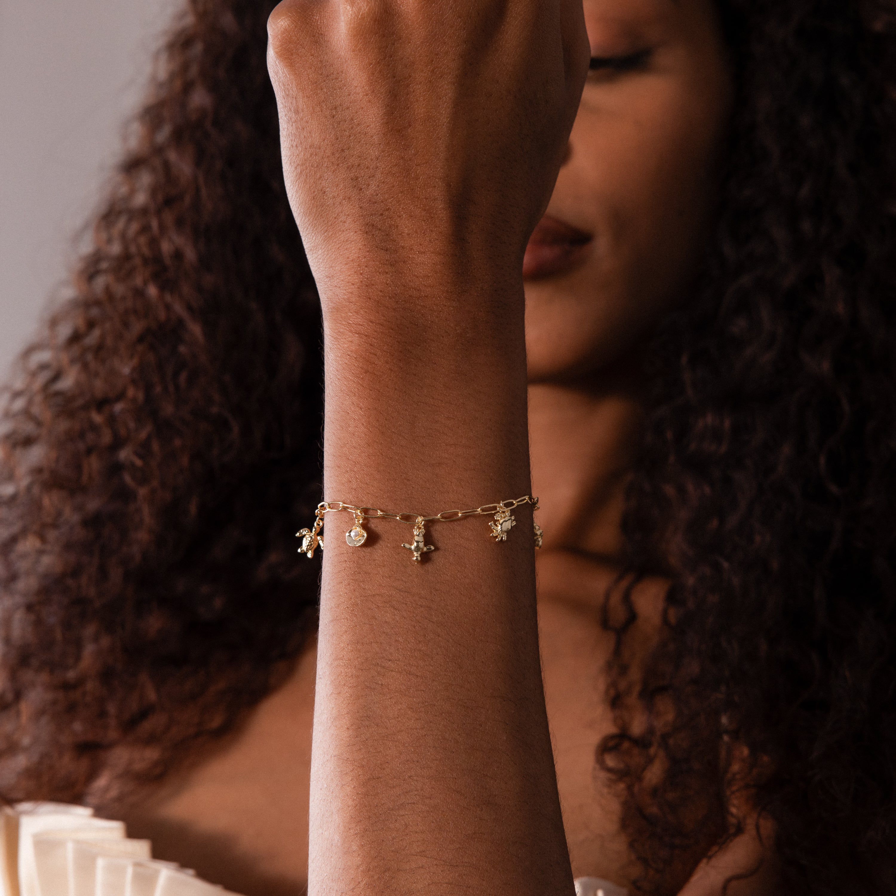 A woman with curly hair poses near a neutral background, showing off the Seaside Charm Bracelet and its ocean-inspired charms.
