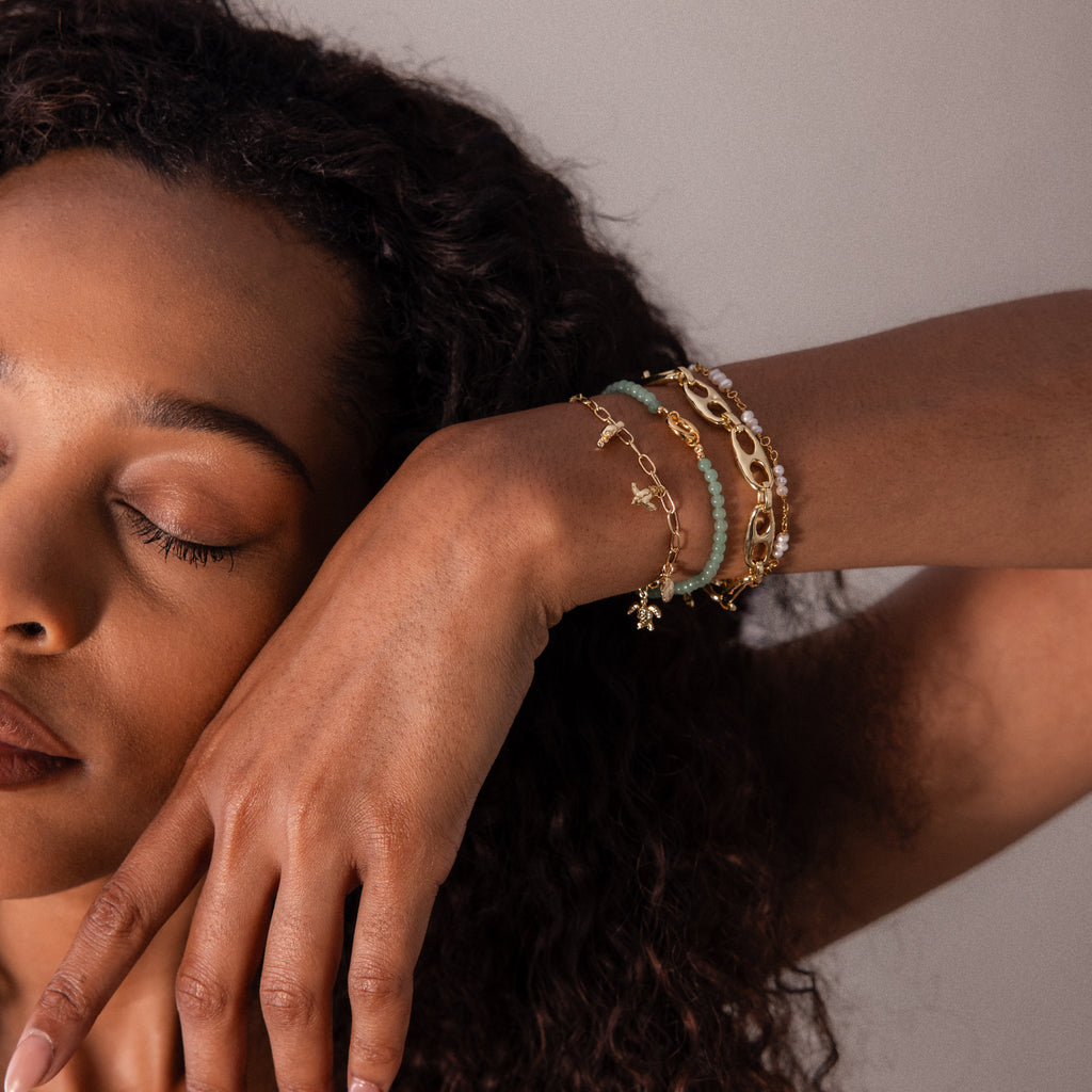 A woman with closed eyes rests her face on her hand, wearing gold and beaded bracelets, including the Seaside Charm Bracelet featuring ocean-inspired charms.