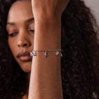 A woman with curly hair raises her wrist to display the Seaside Charm Bracelet in Silver, adorned with ocean-inspired charms, while her face remains softly out of focus.