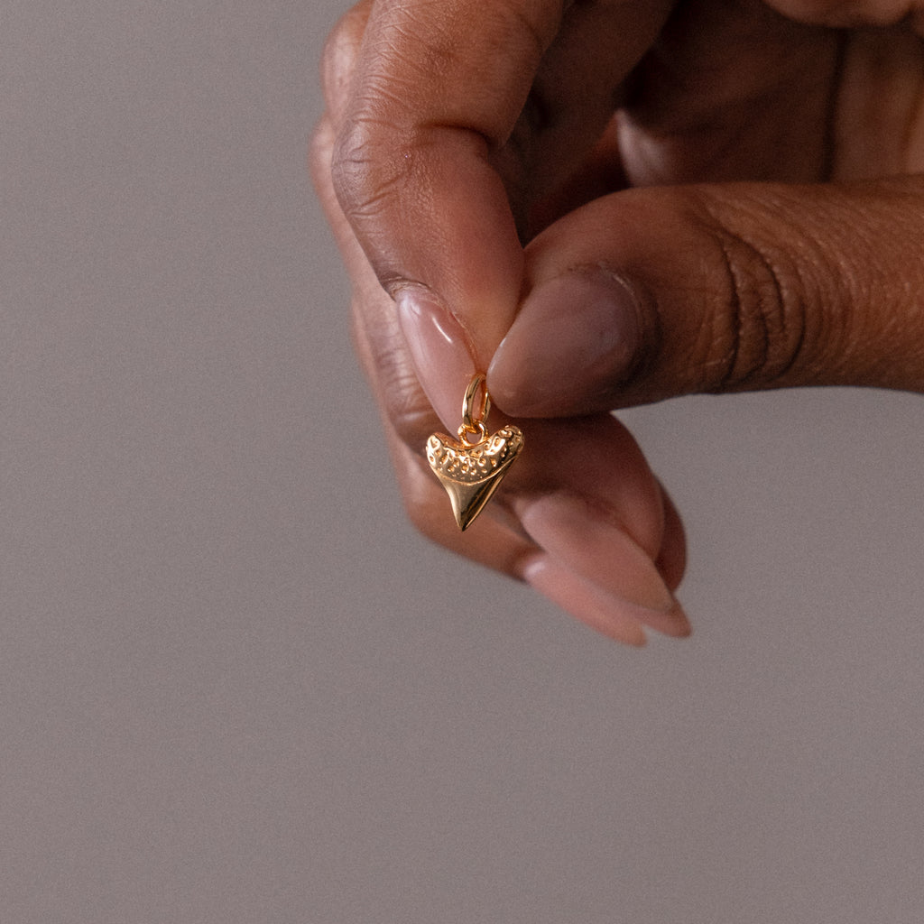 A hand holds the Dainty Shark Tooth Charm—a small gold pendant with a row of crystals on top, exuding coastal charm and perfect for adding beach jewelry elegance to any look.