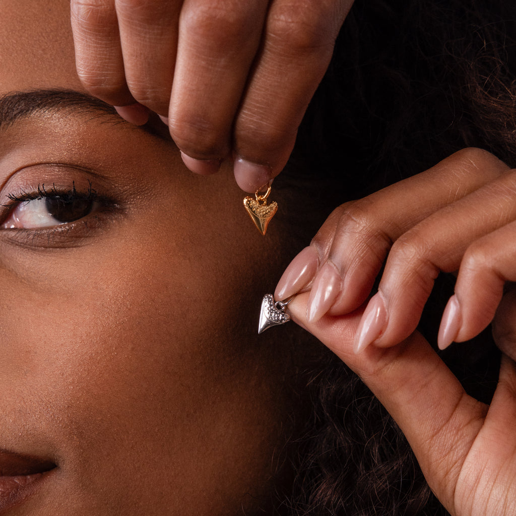 A person holds gold and silver Dainty Shark Tooth Charms near their face, showcasing both pieces and highlighting the coastal charm often found in beach jewelry.