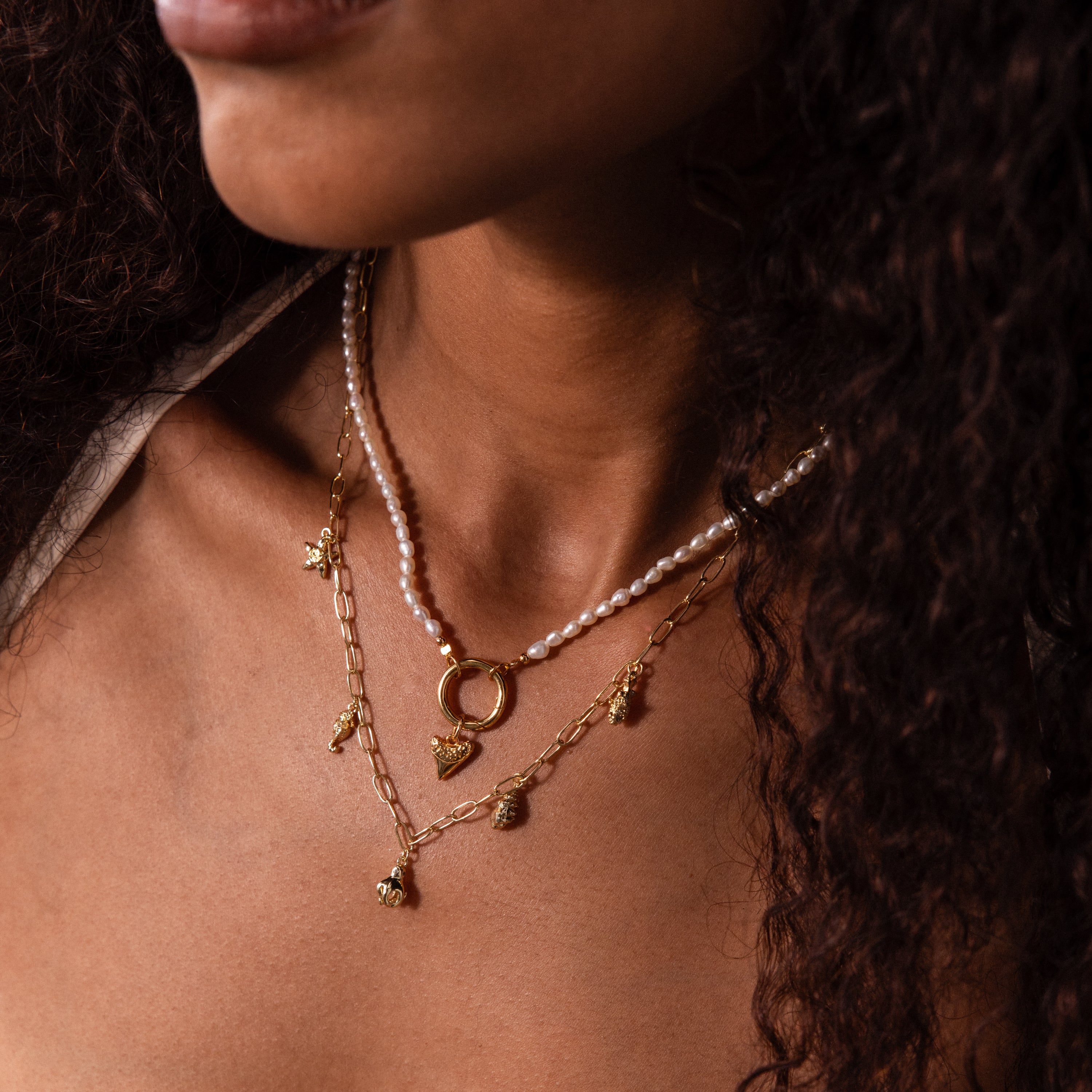 A woman wears layered gold and pearl necklaces featuring the Dainty Shark Tooth Charm; only her neck, chin, and curly hair show, evoking effortless coastal style.