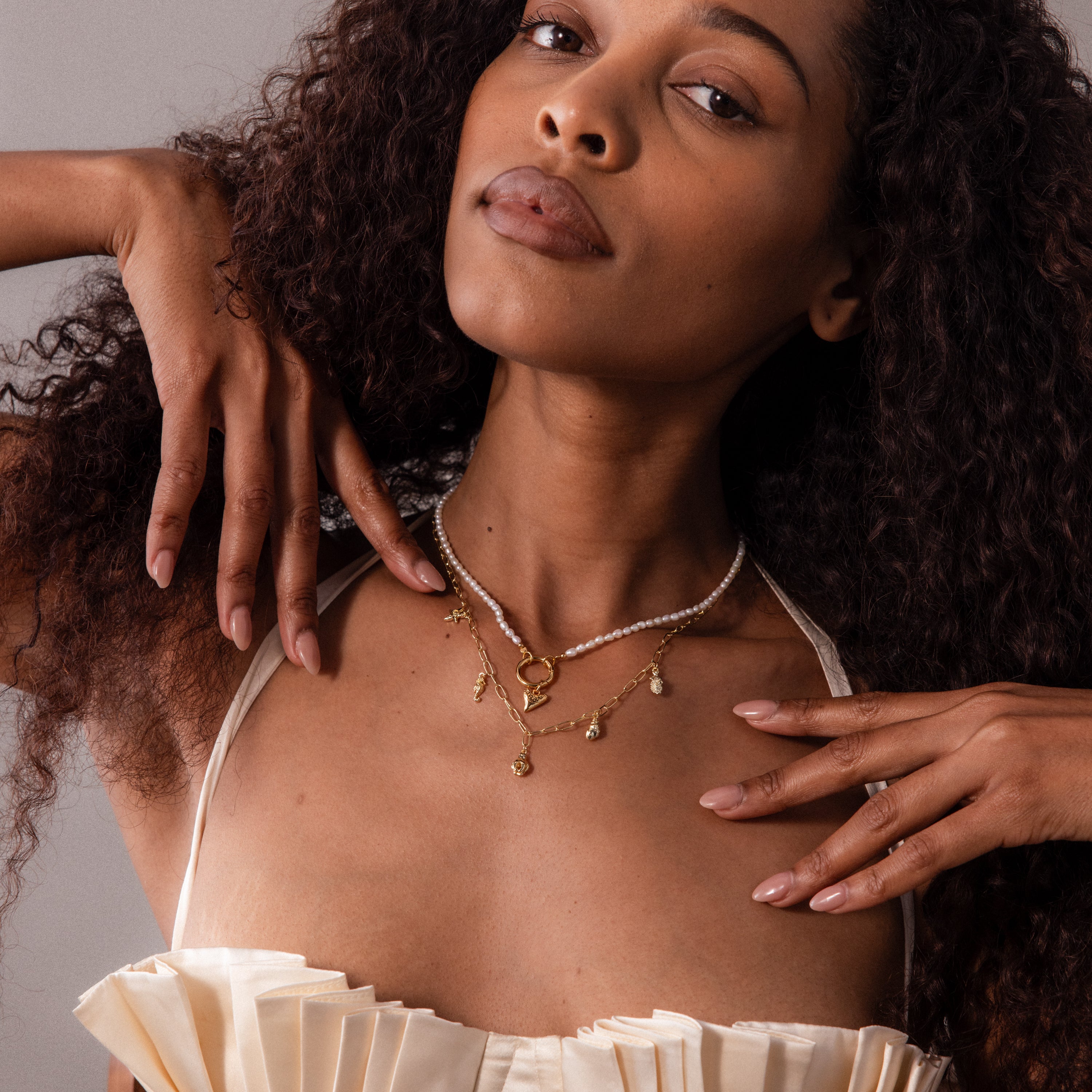 A woman with curly hair wears a cream pleated top and layered gold necklaces, featuring the Dainty Shark Tooth Charm, posing with hands near her shoulders for effortless coastal charm.