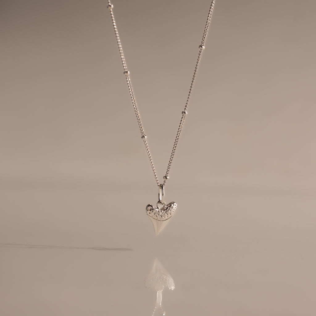 The Shark Tooth Necklace in Silver, displayed against a soft beige background, adds subtle coastal charm and beachside style to any look.