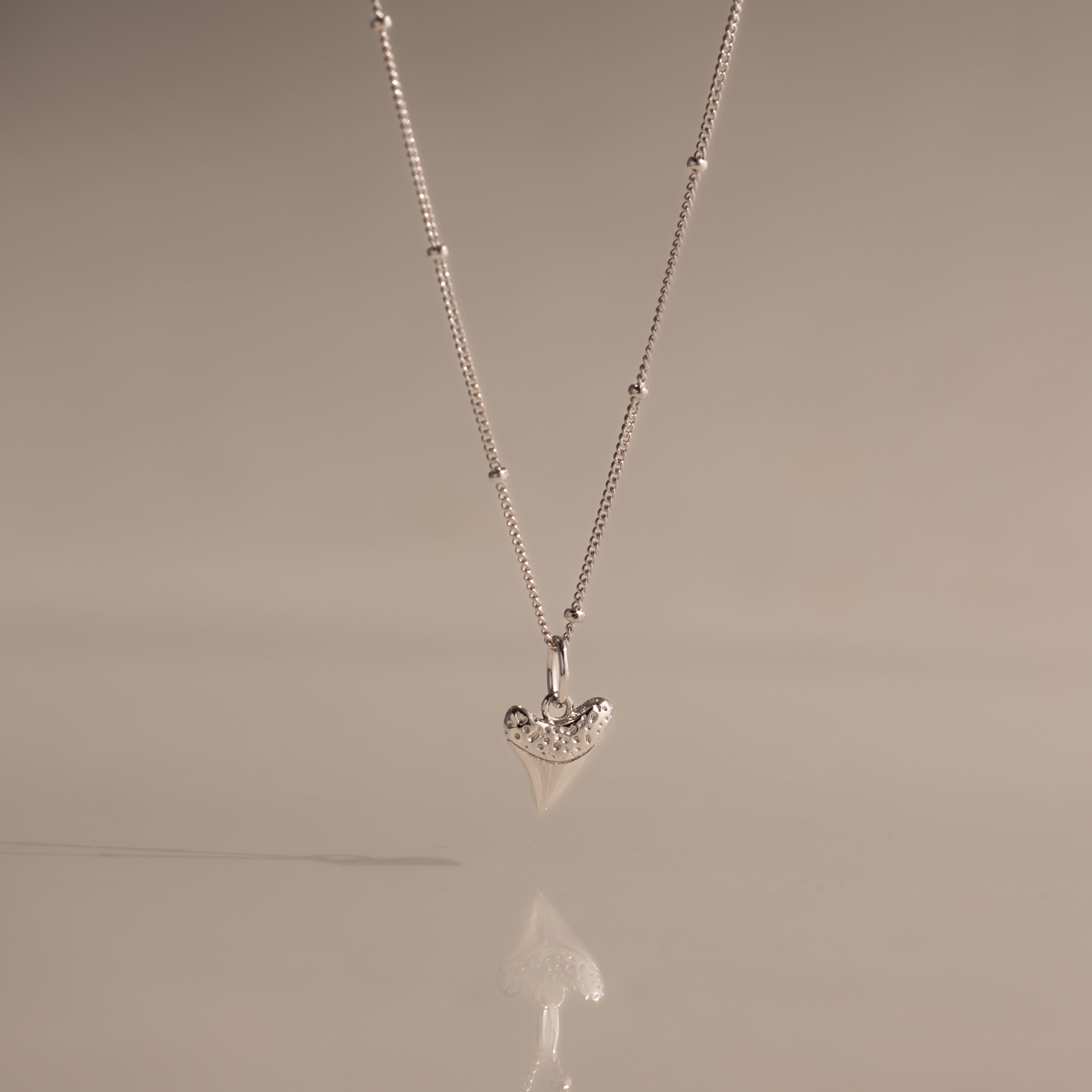 The Shark Tooth Necklace in Silver, displayed against a soft beige background, adds subtle coastal charm and beachside style to any look.