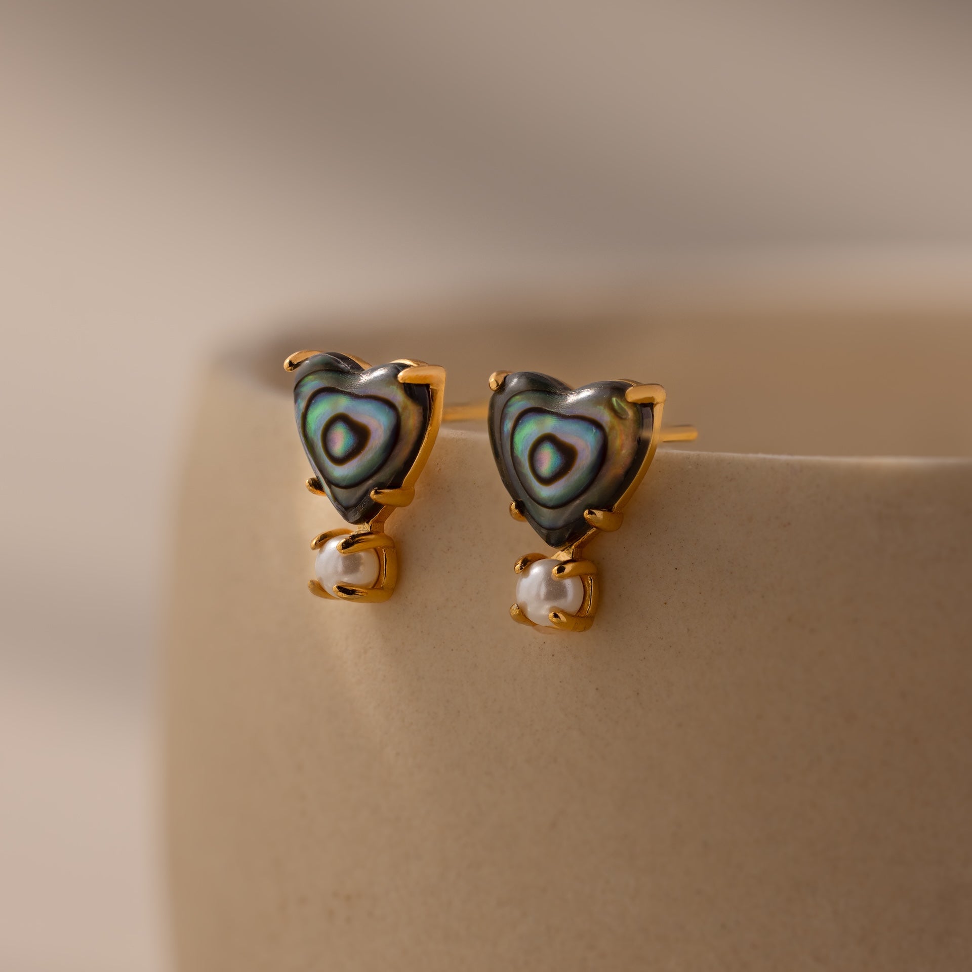 Abalone Pearl Heart Studs in 18K Gold featuring abalone inlay and delicate pearl accents, perfect coastal jewelry shown on a beige ceramic surface.