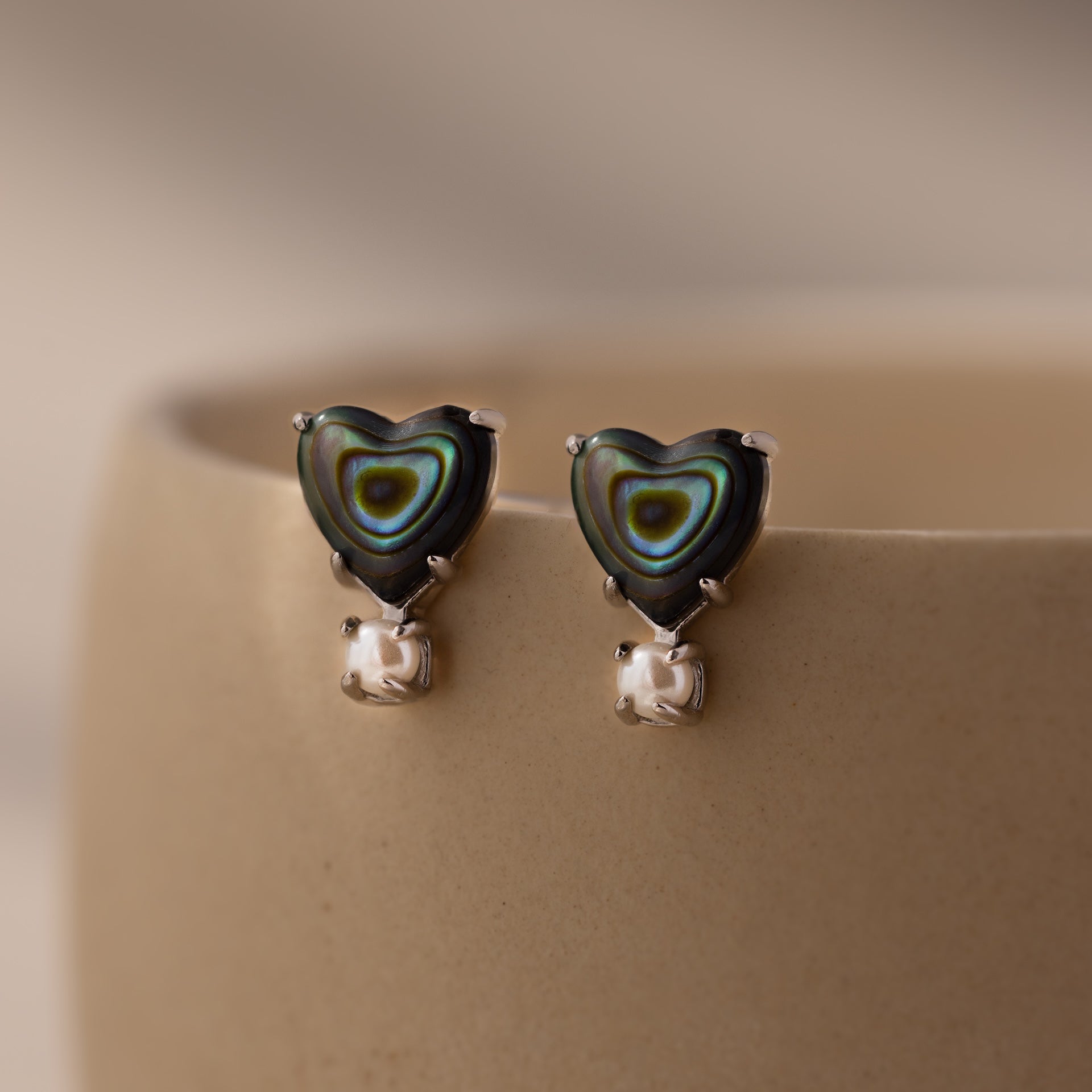 Abalone Pearl Heart Studs in Silver, displayed on a beige ceramic cup, are perfect for lovers of coastal jewelry.
