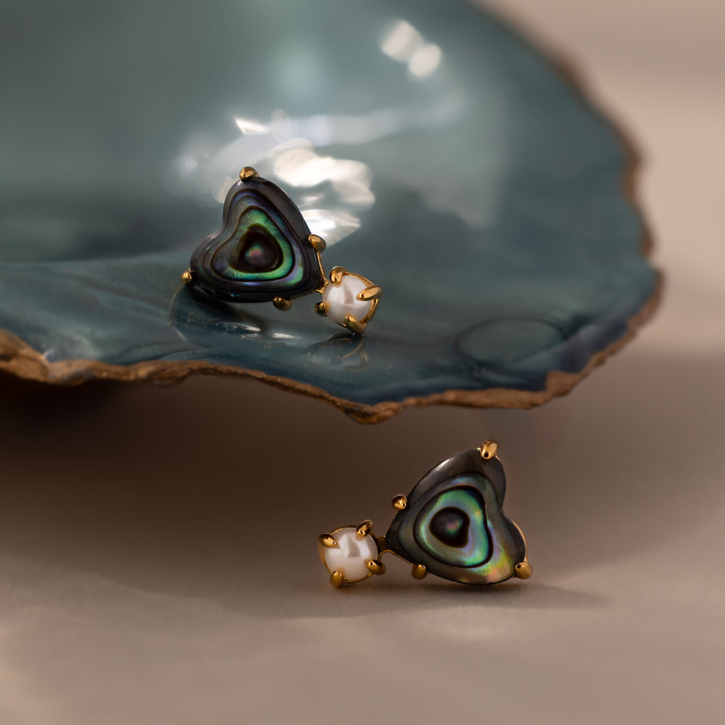 Abalone Pearl Heart Studs in 18K Gold rest elegantly on a blue ceramic dish, their heart-shaped design and gold accents evoking coastal jewelry charm.