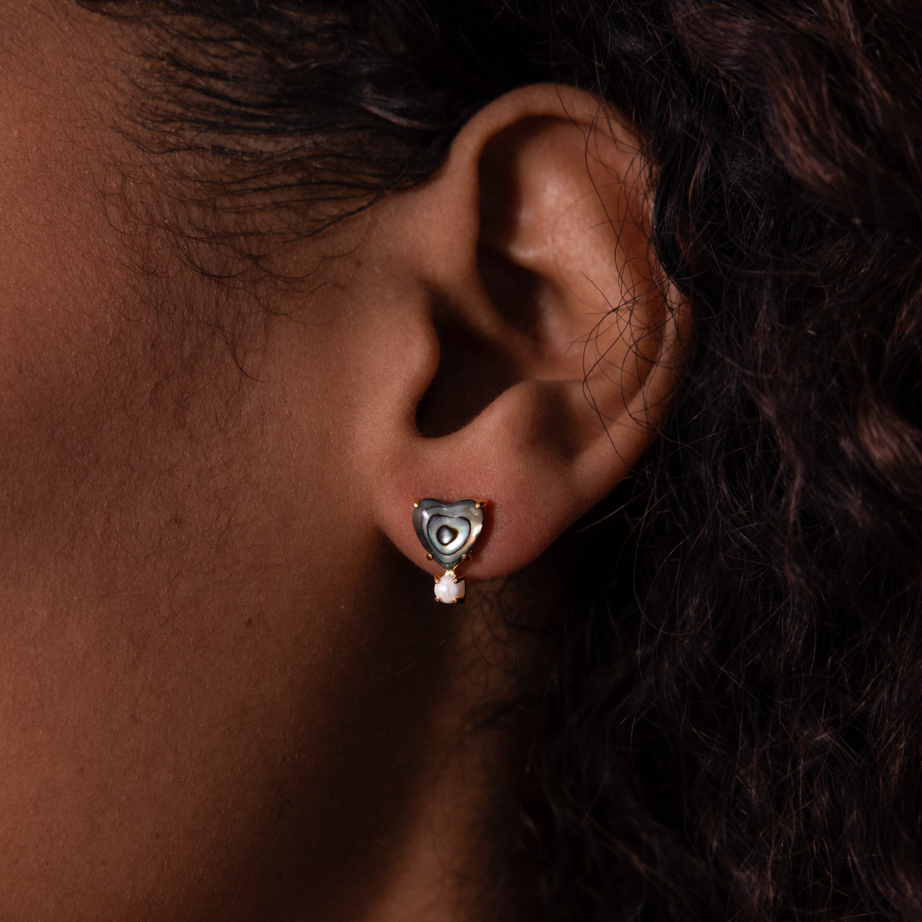 Close-up of an ear wearing Abalone Pearl Heart Studs in 18K Gold, featuring a heart-shaped stud with a small dangling gem for a charming coastal look.