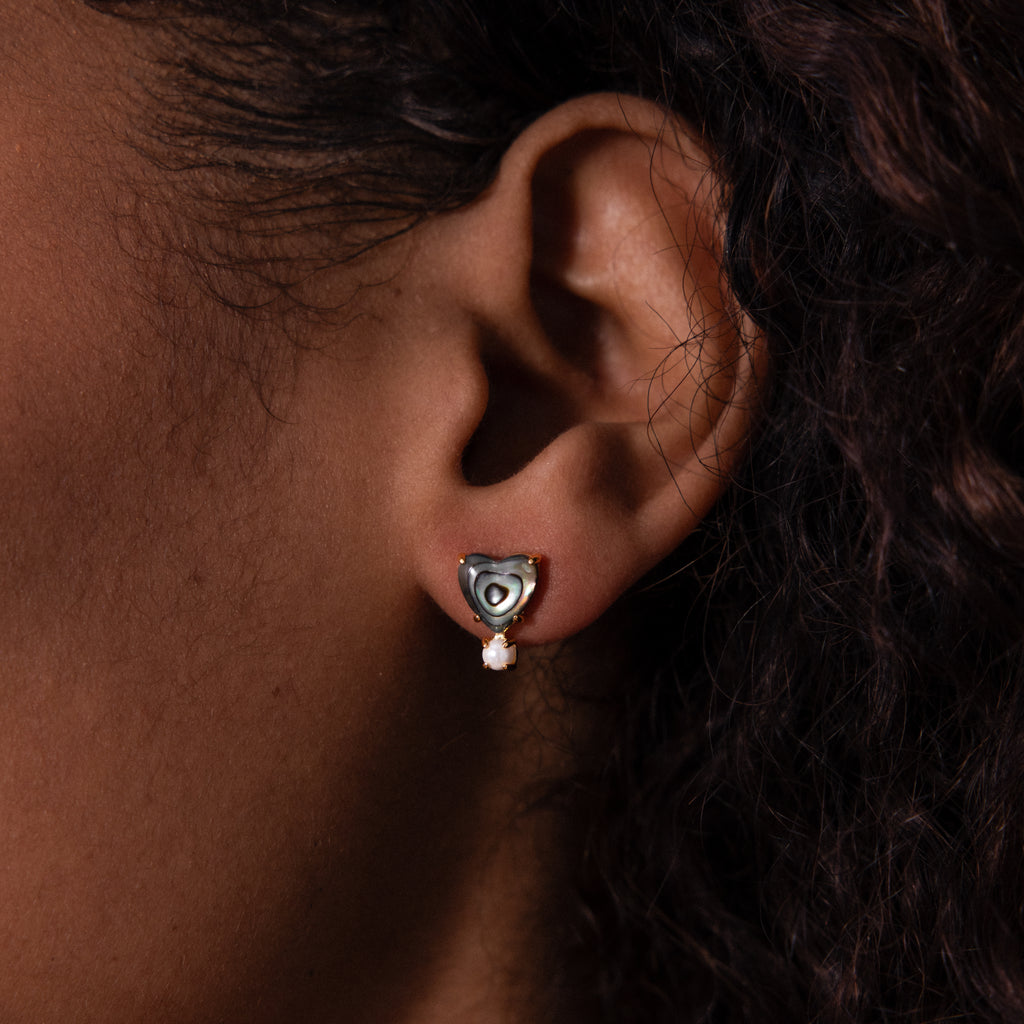 A close-up of an ear wearing Abalone Pearl Heart Studs—heart-shaped silver studs with a dangling pearl—captures the delicate beauty of coastal jewelry.