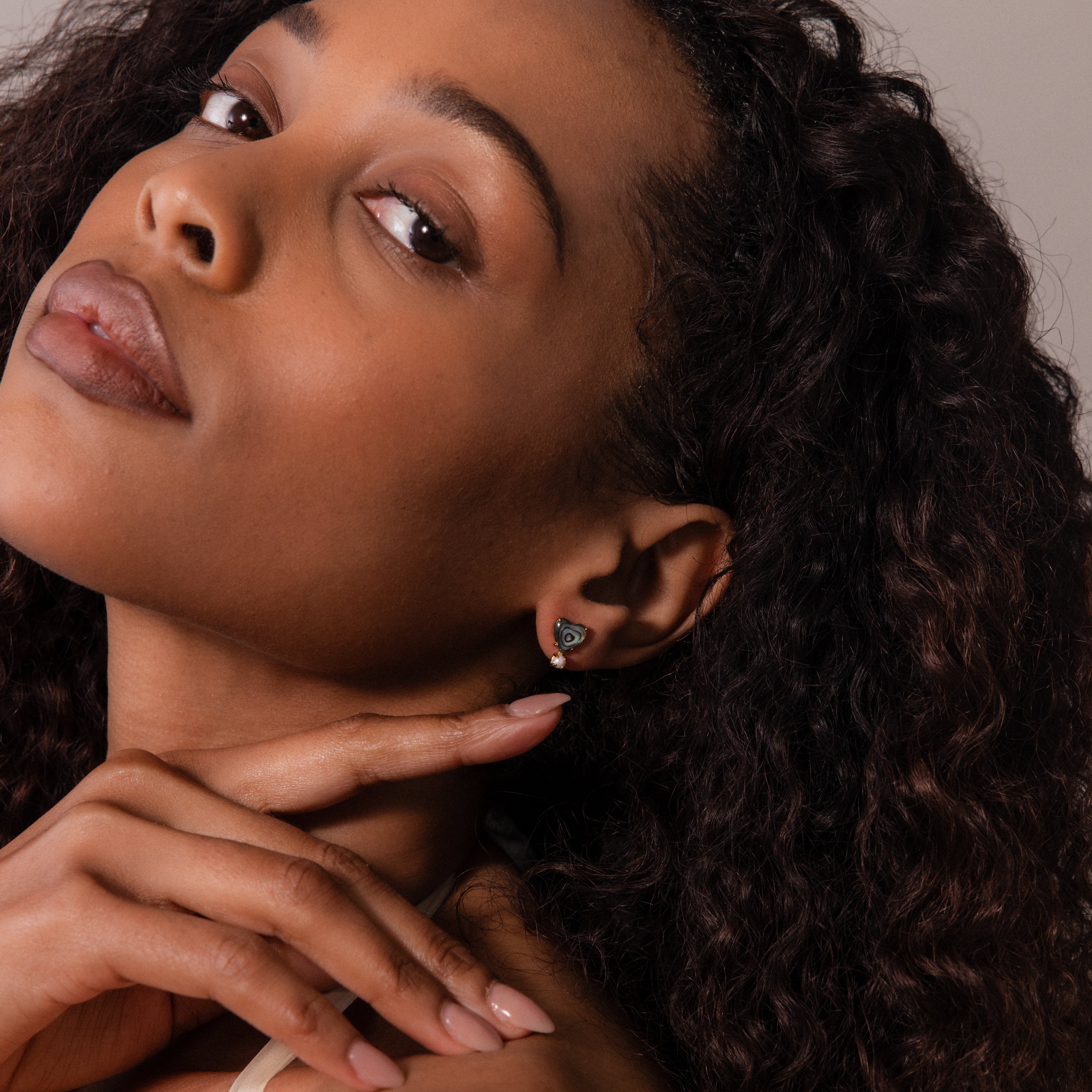 Woman with curly hair tilts her head, showcasing Abalone Pearl Heart Studs in 18K Gold while resting her hands under her chin.