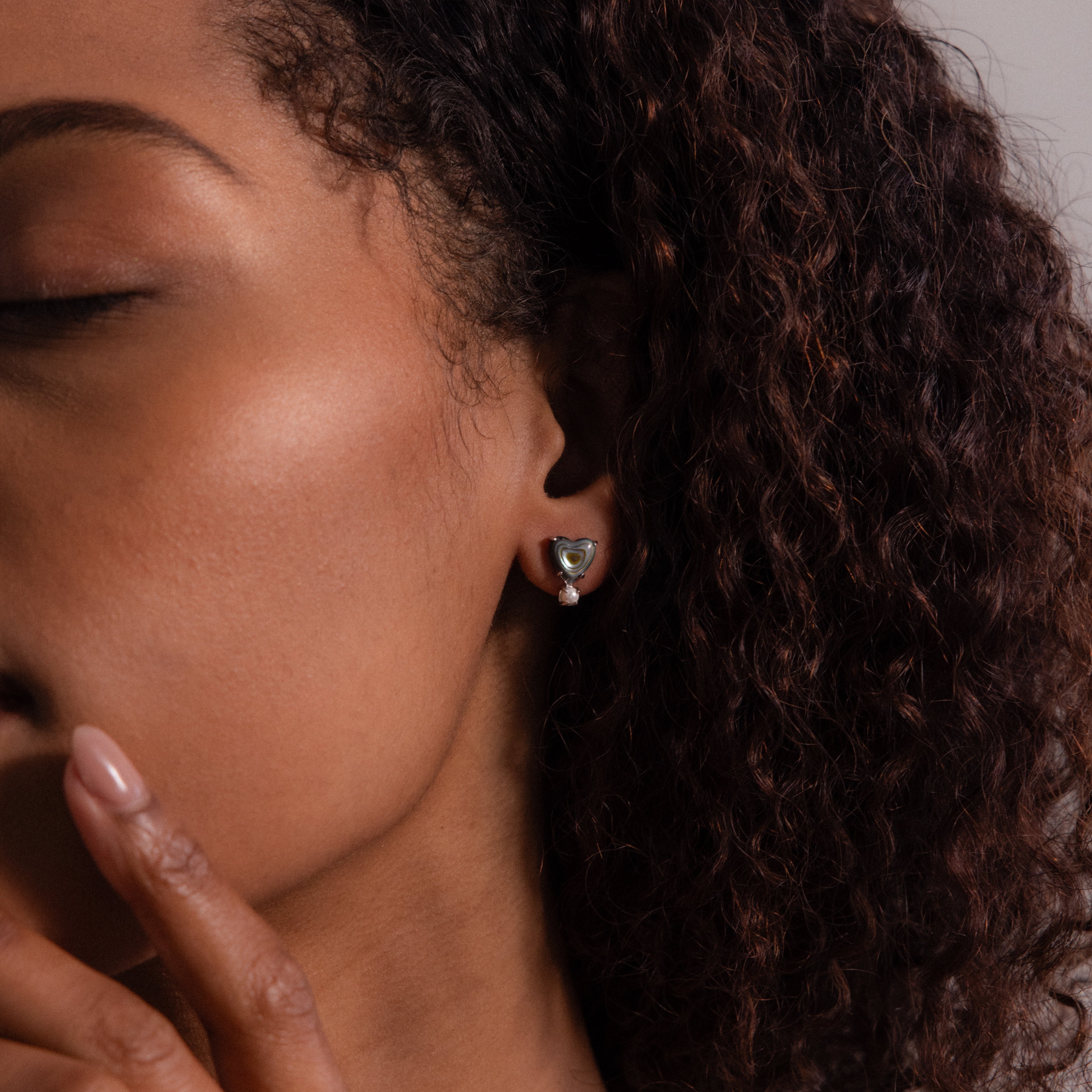 A woman with curly hair, eyes closed, gently touches her face while wearing Abalone Pearl Heart Studs in Silver, embodying the serene elegance of coastal jewelry.