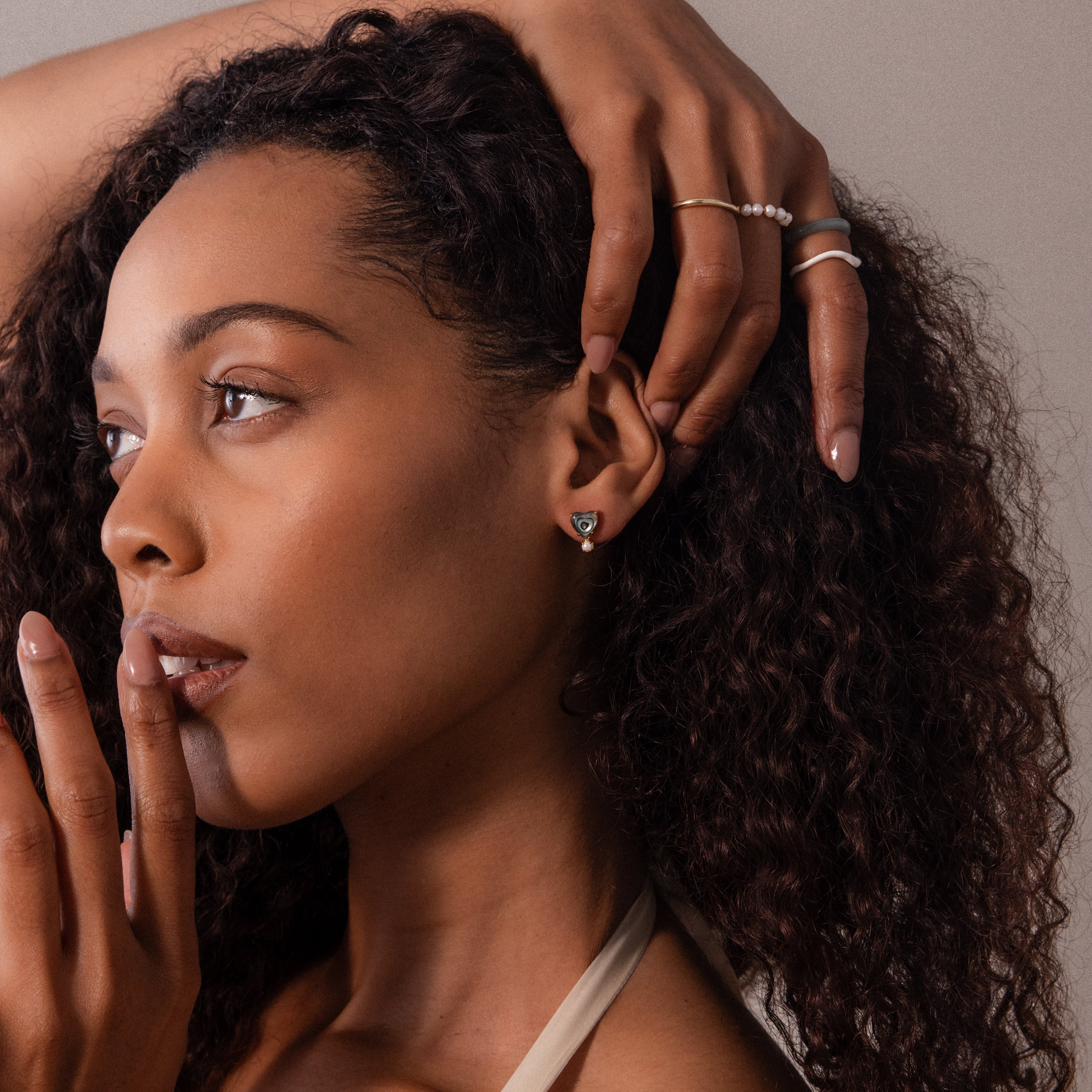 Woman with curly hair wears Abalone Pearl Heart Studs in 18K Gold, rings, and coastal jewelry, touching her face with one hand.