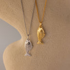 The Fish Pendant Necklace in 18K Gold is displayed on a chain against a textured beige surface, perfect for ocean-inspired style or layering.
