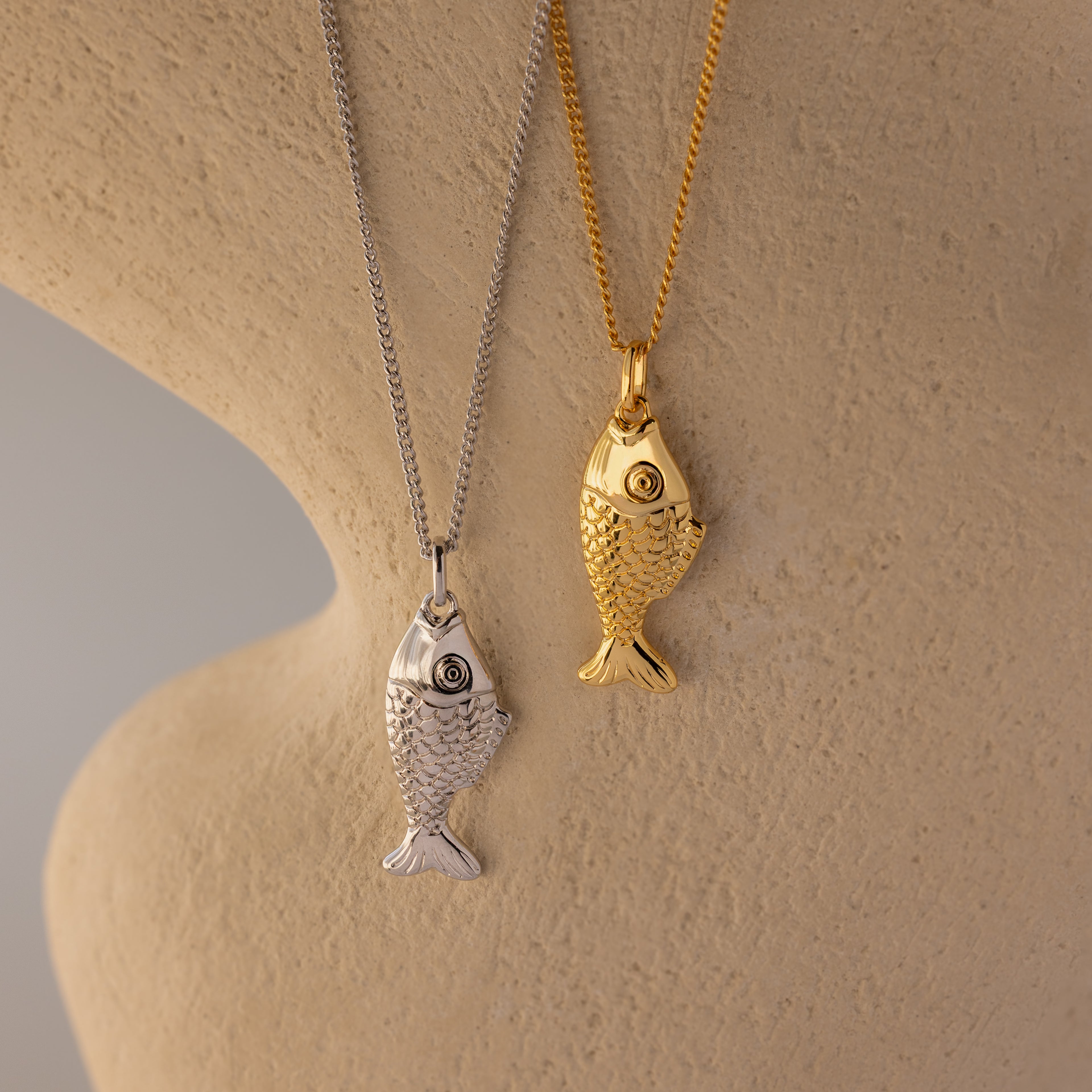 The Fish Pendant Necklace in 18K Gold is displayed on a chain against a textured beige surface, perfect for ocean-inspired style or layering.
