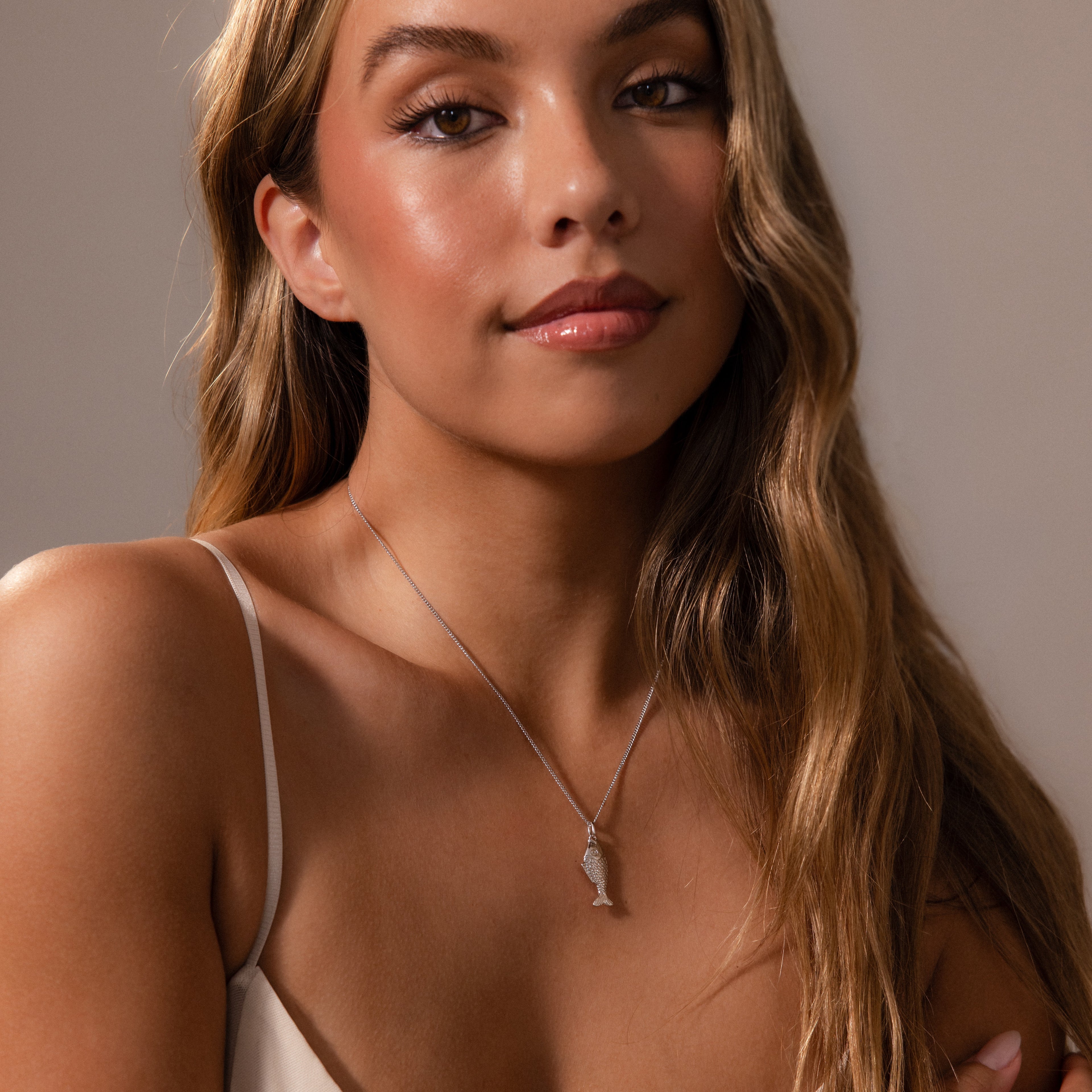 Woman with long wavy hair wears a light beige camisole and the Fish Pendant Necklace in Silver, looking at the camera.