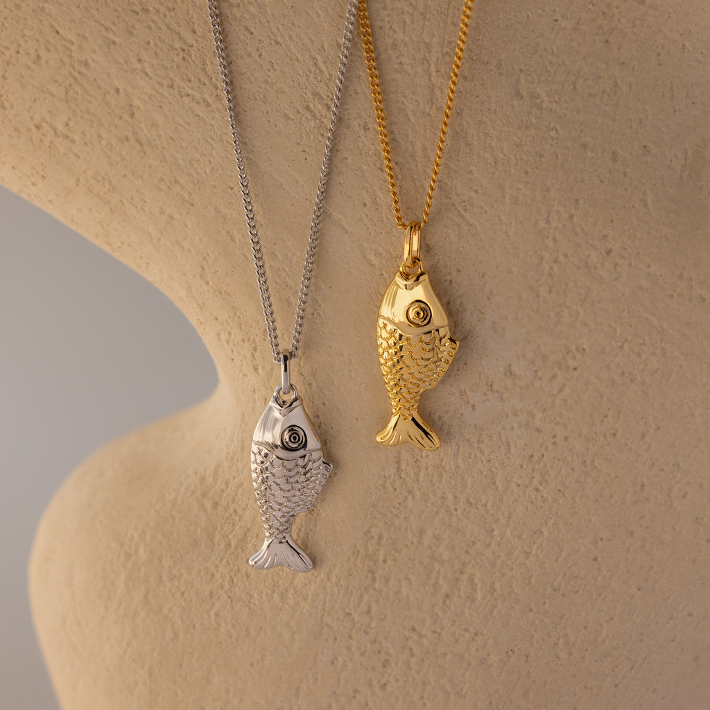 The Fish Pendant Necklace in Silver features a fish-shaped pendant for an elegant, ocean-inspired look, displayed on a beige textured surface.