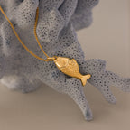 The Fish Pendant Necklace in 18K Gold drapes over textured light gray coral—an ocean-inspired piece perfect for effortless layering.