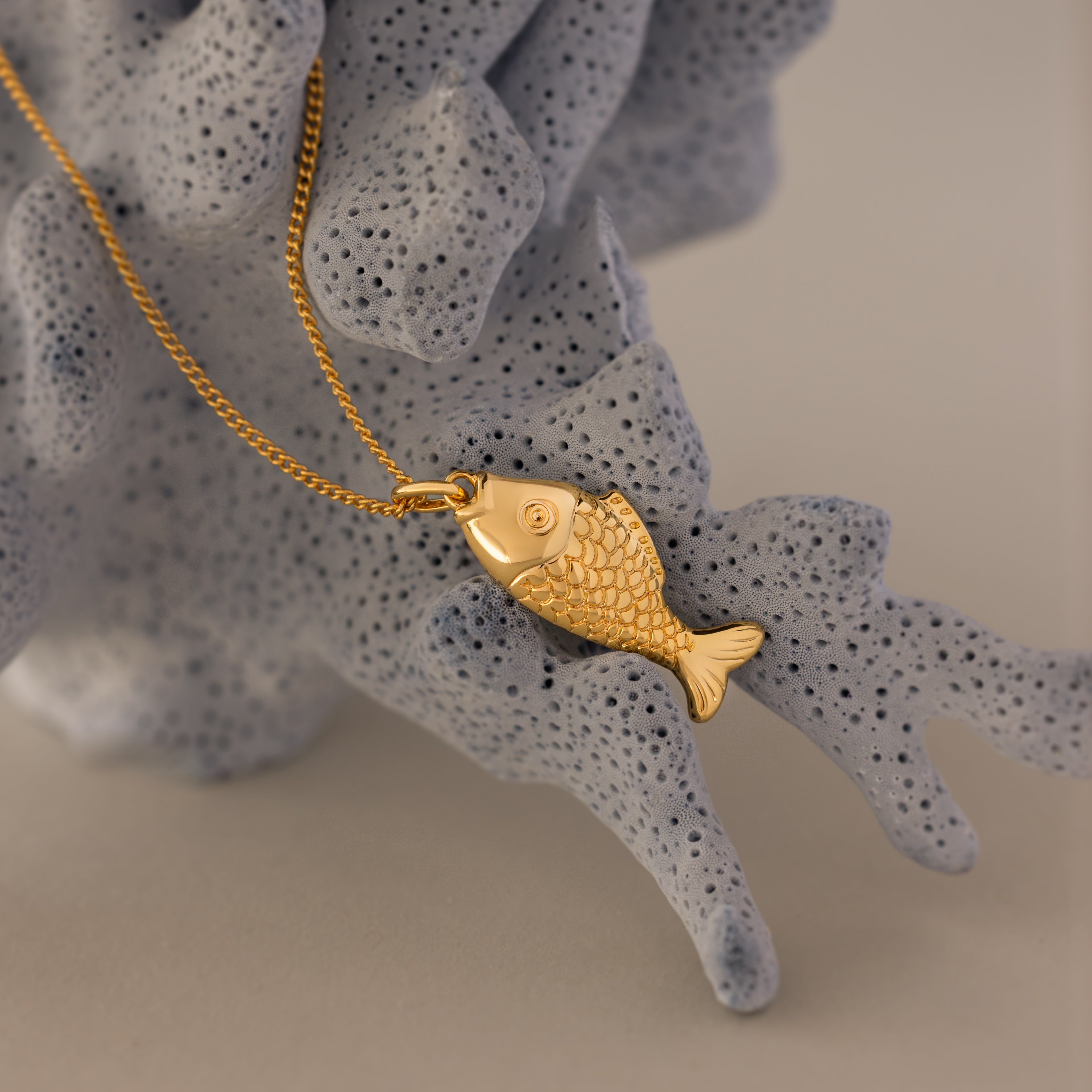The Fish Pendant Necklace in 18K Gold drapes over textured light gray coral—an ocean-inspired piece perfect for effortless layering.