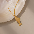The Fish Pendant Necklace in 18K Gold rests on a white seashell background, making it an ideal delicate layering necklace for every beach lover.