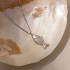 The Fish Pendant Necklace in Silver is displayed on an iridescent seashell in soft natural light, perfect for layering and adding a stylish, beachy touch to any look.