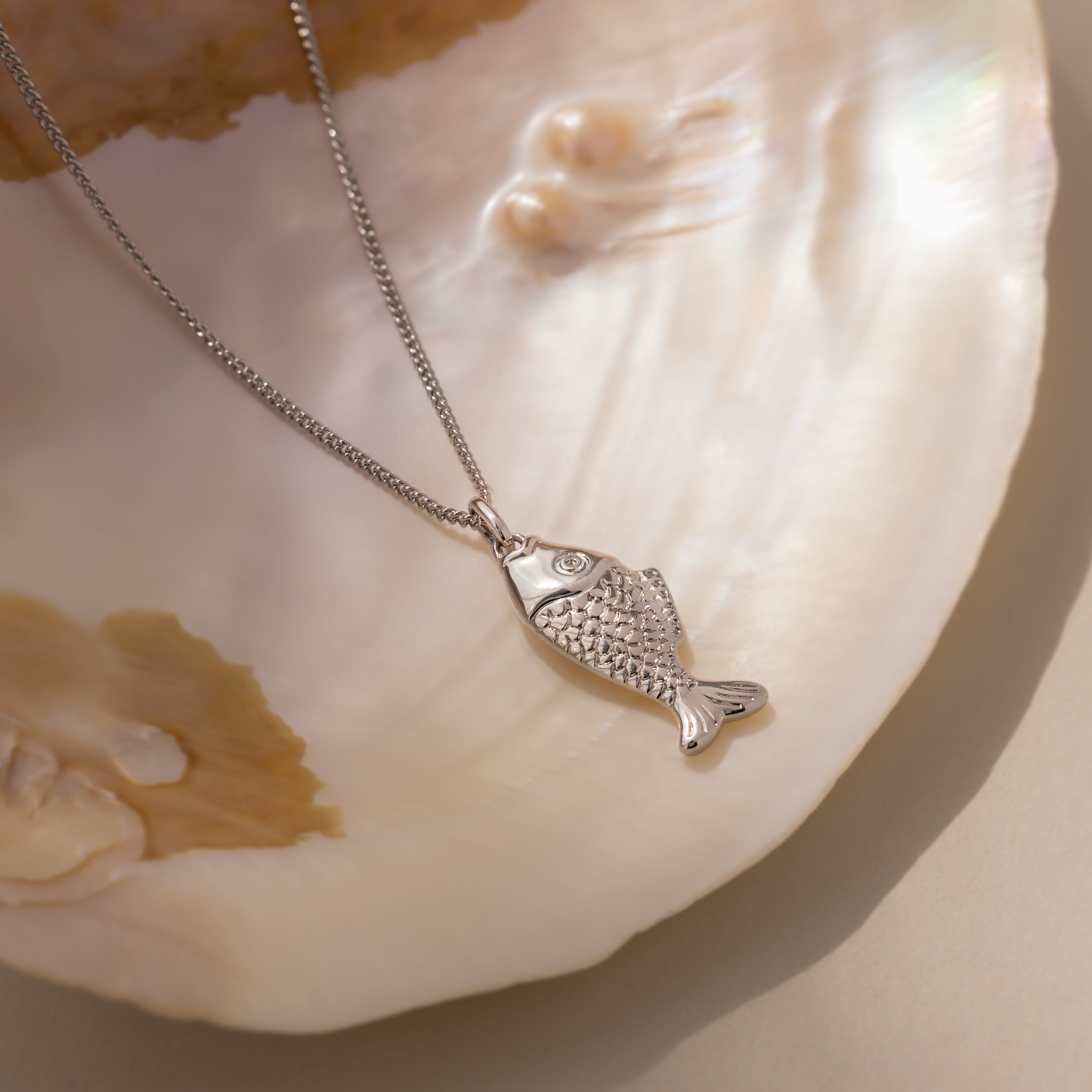 The Fish Pendant Necklace in Silver is displayed on an iridescent seashell in soft natural light, perfect for layering and adding a stylish, beachy touch to any look.