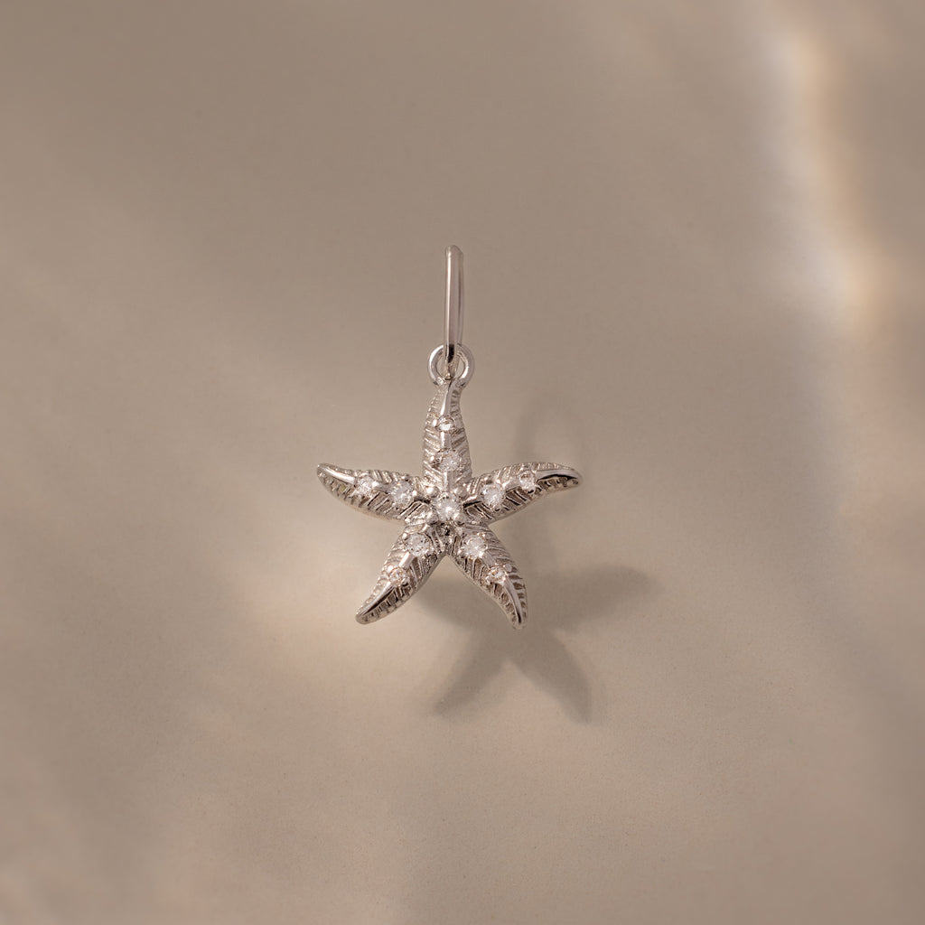 The Pave Starfish Charm in Silver is displayed on a smooth beige background with soft light reflections, highlighting the elegant coastal charm of this starfish jewelry.