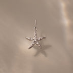 The Pave Starfish Charm in Silver is displayed on a smooth beige background with soft light reflections, highlighting the elegant coastal charm of this starfish jewelry.