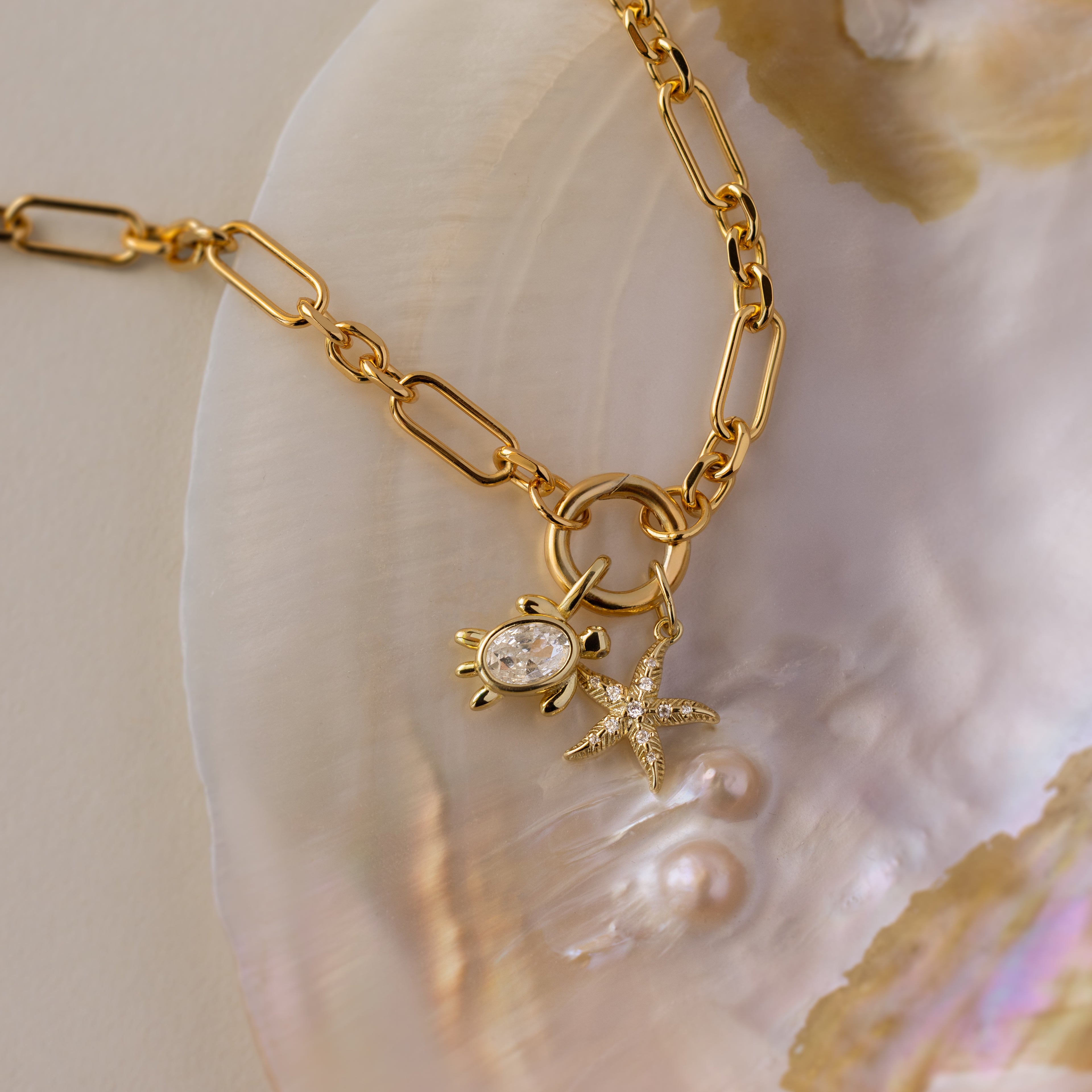 Add coastal charm to your look with the Pave Starfish Charm—a gold chain necklace with crystal accents, displayed on a pearlescent seashell background.