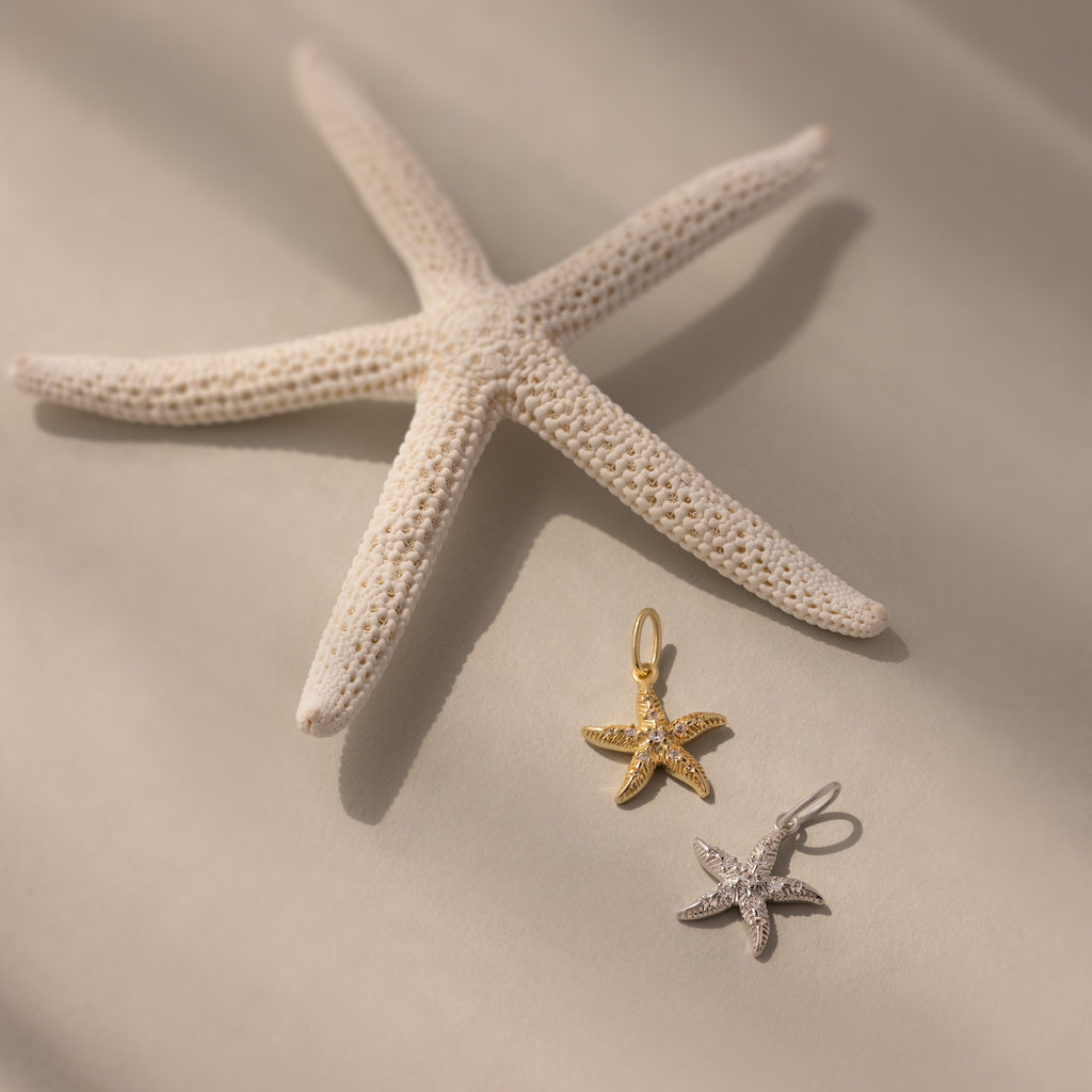 A white starfish highlights the Pave Starfish Charm in gold and silver on a soft, neutral background, showcasing its coastal elegance.