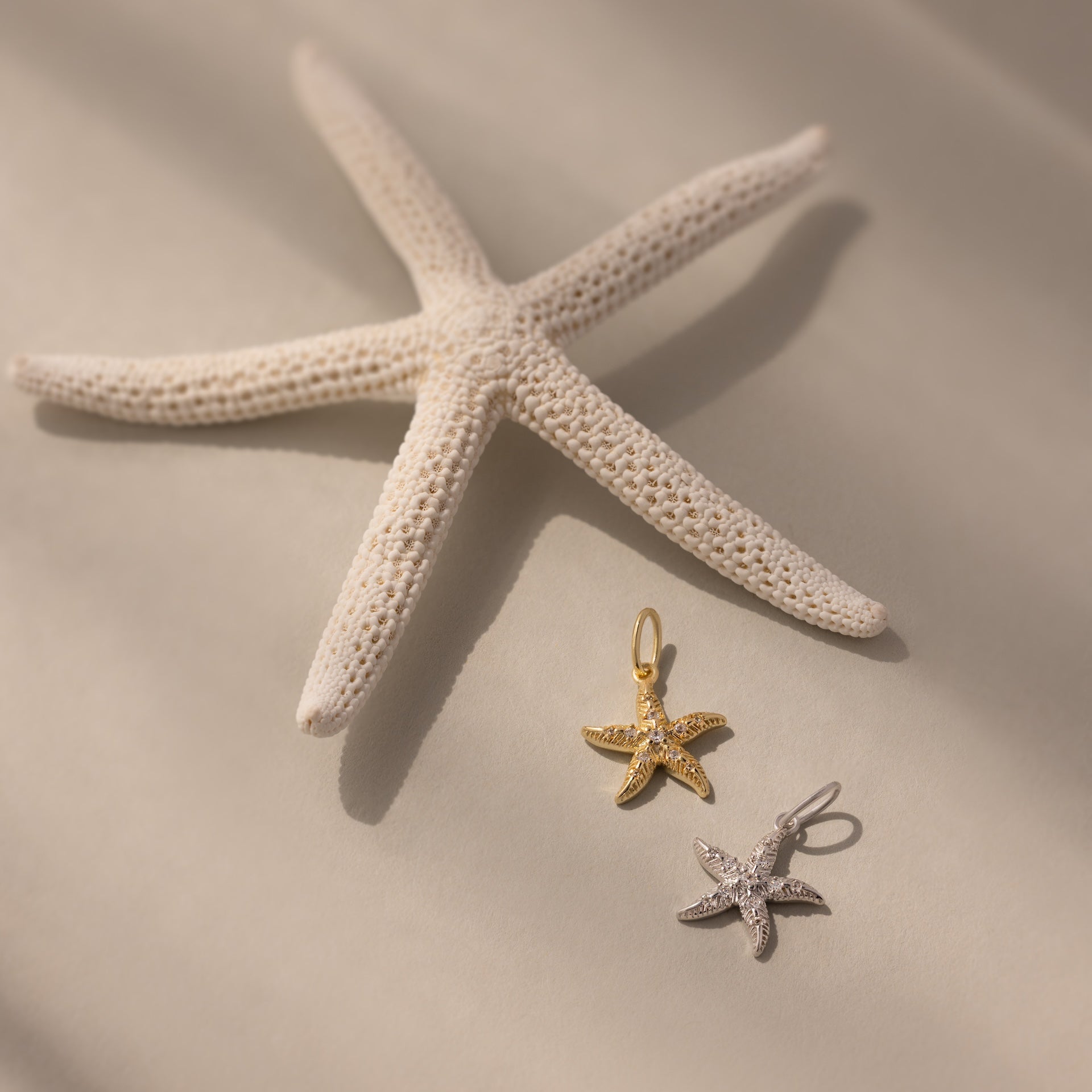 A white starfish highlights the Pave Starfish Charm in gold and silver on a soft, neutral background, showcasing its coastal elegance.