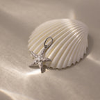 The Pave Starfish Charm shines atop a white seashell against a soft, shimmering background, capturing genuine coastal charm.